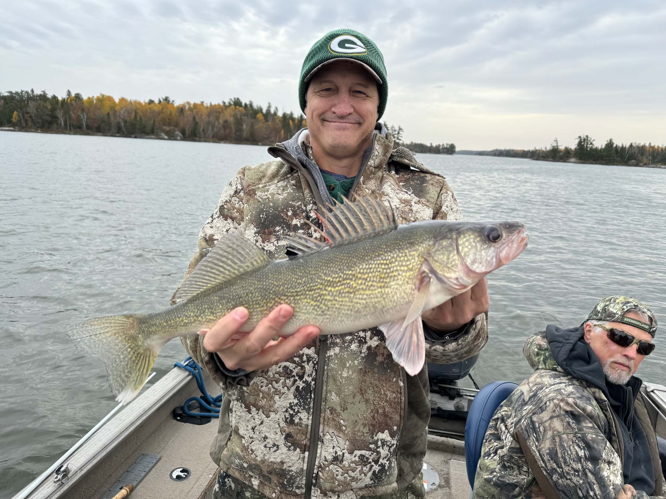 A man in camouflage clothing and a Green Bay Packers beanie holding a large fish on a boat in a body of water with a tree-lined shoreline in the background.