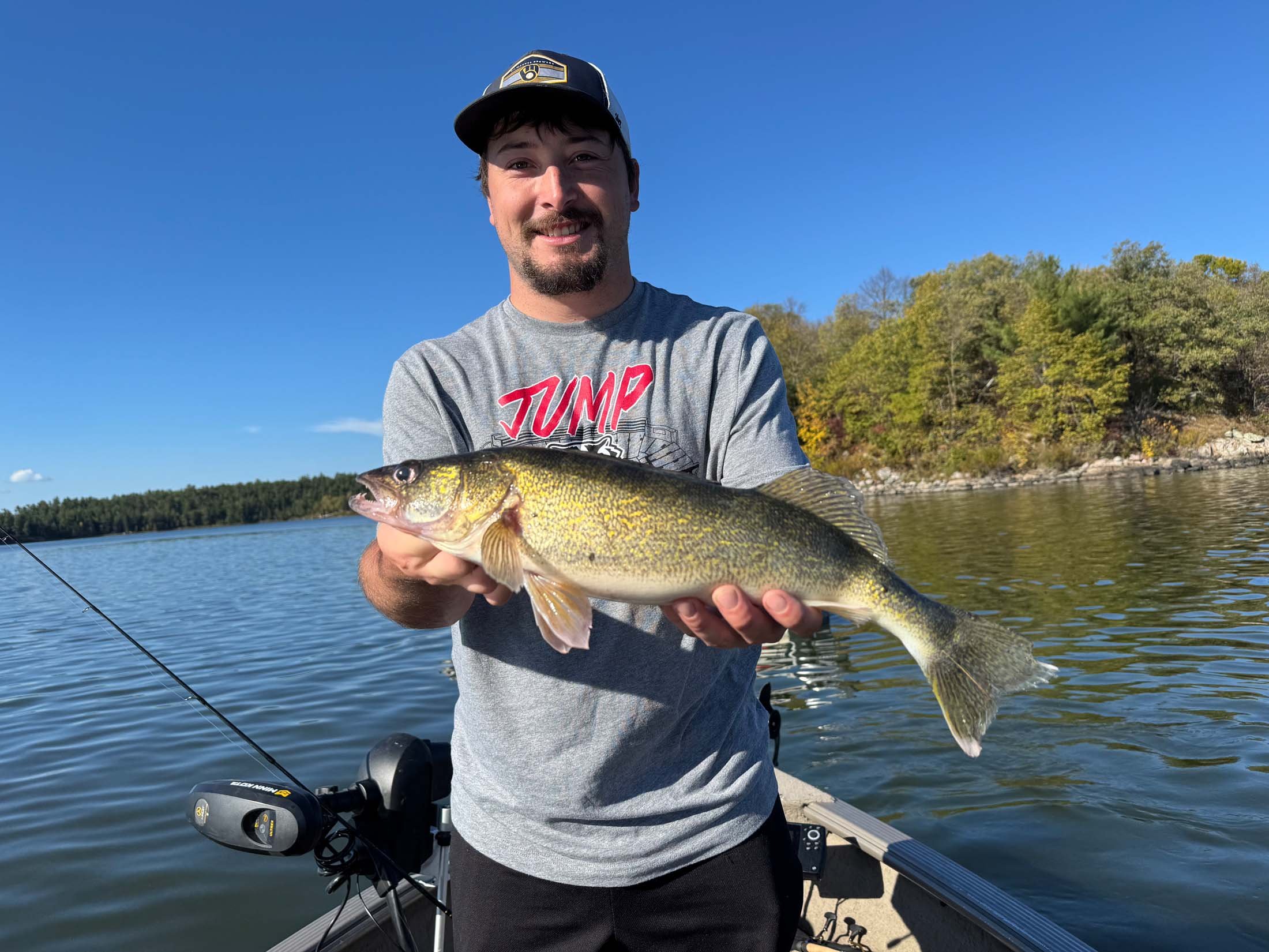 A man smiling and holding a large fish he caught while on a boat in a lake, with trees and a clear sky in the background.