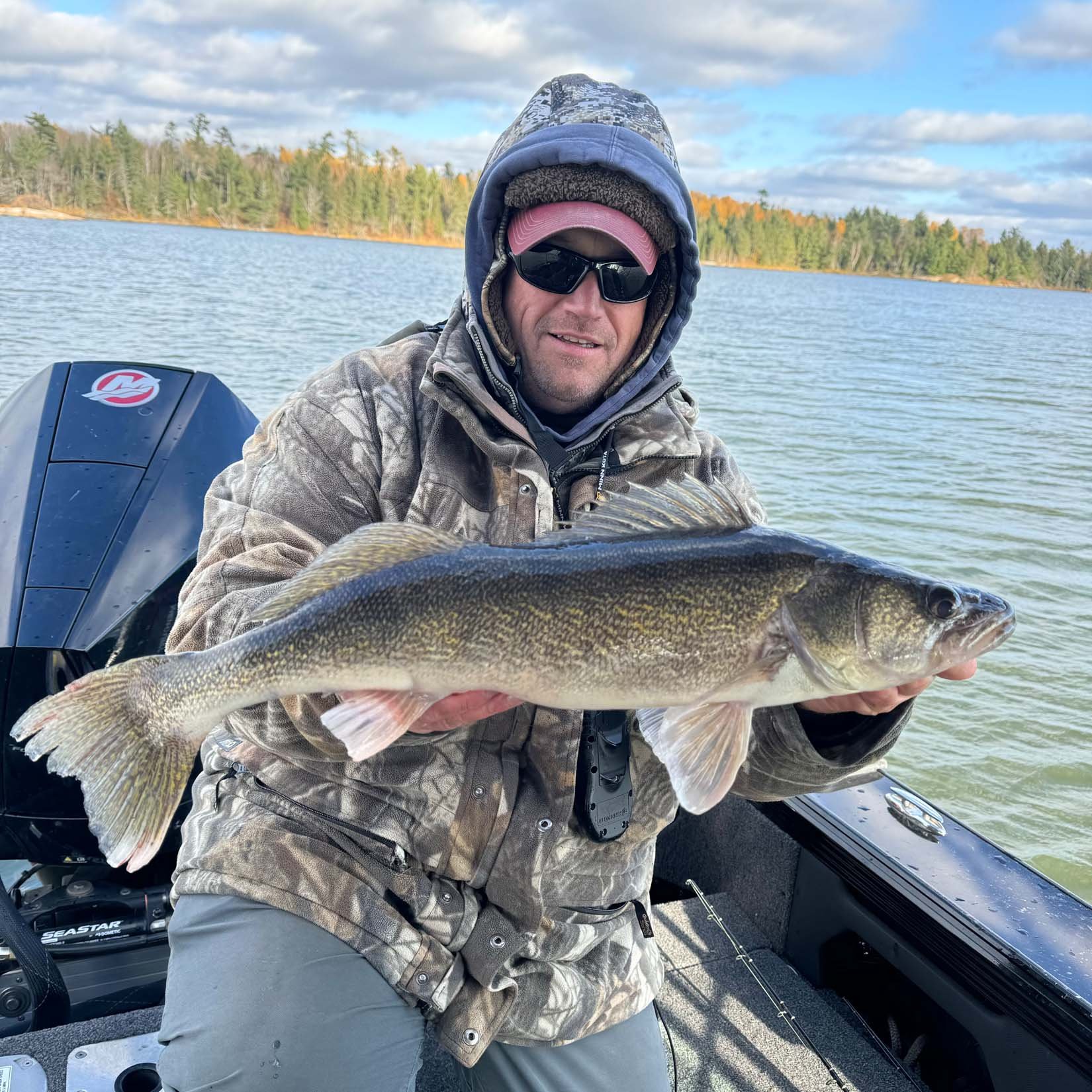 Man in camouflage jacket, sunglasses, and pink cap on a boat holding a large fish with water and trees in the background.