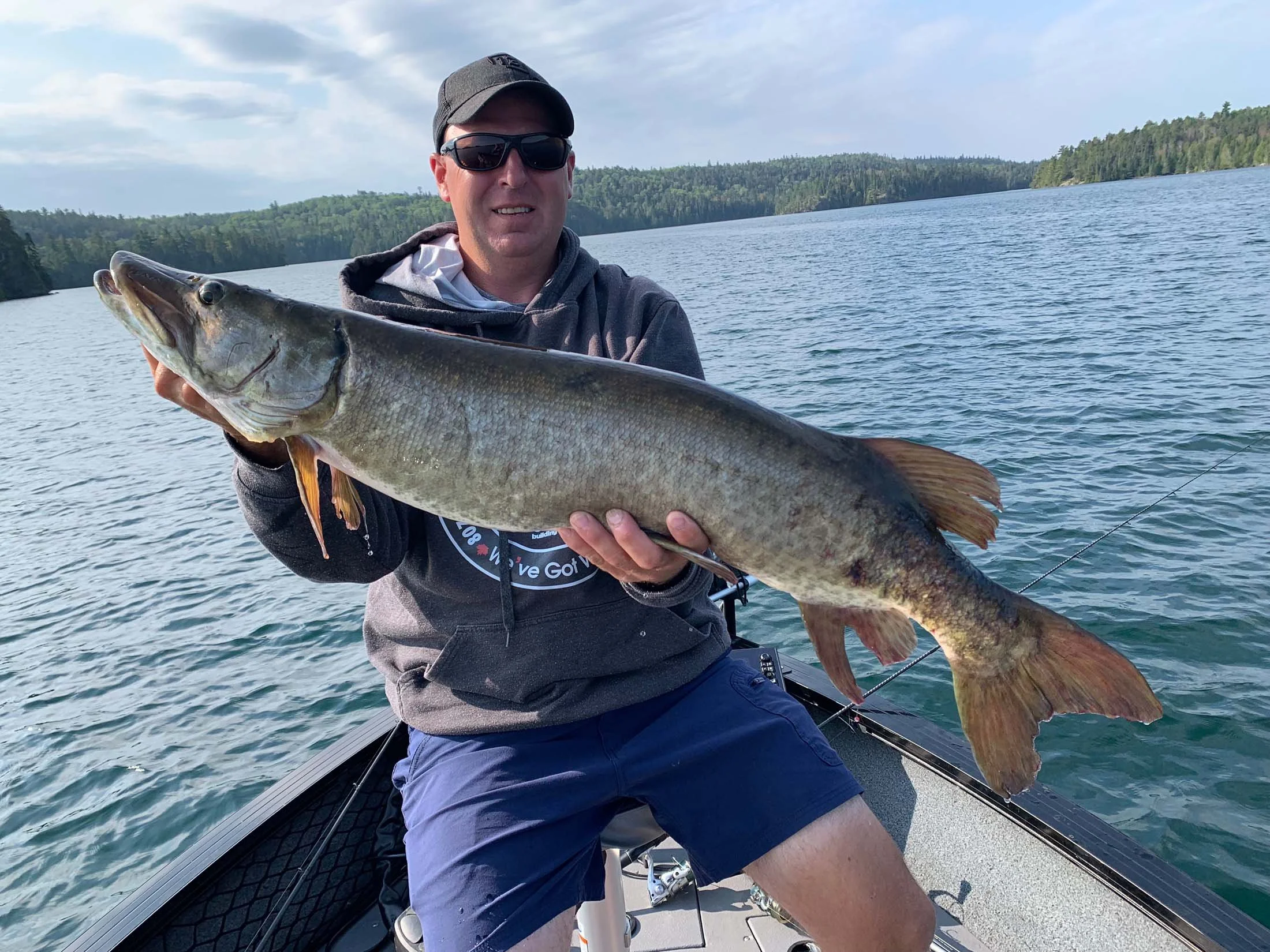 A man wearing sunglasses, a black cap, and a gray hoodie holding a large fish on a boat in a lake with forested hills in the background.