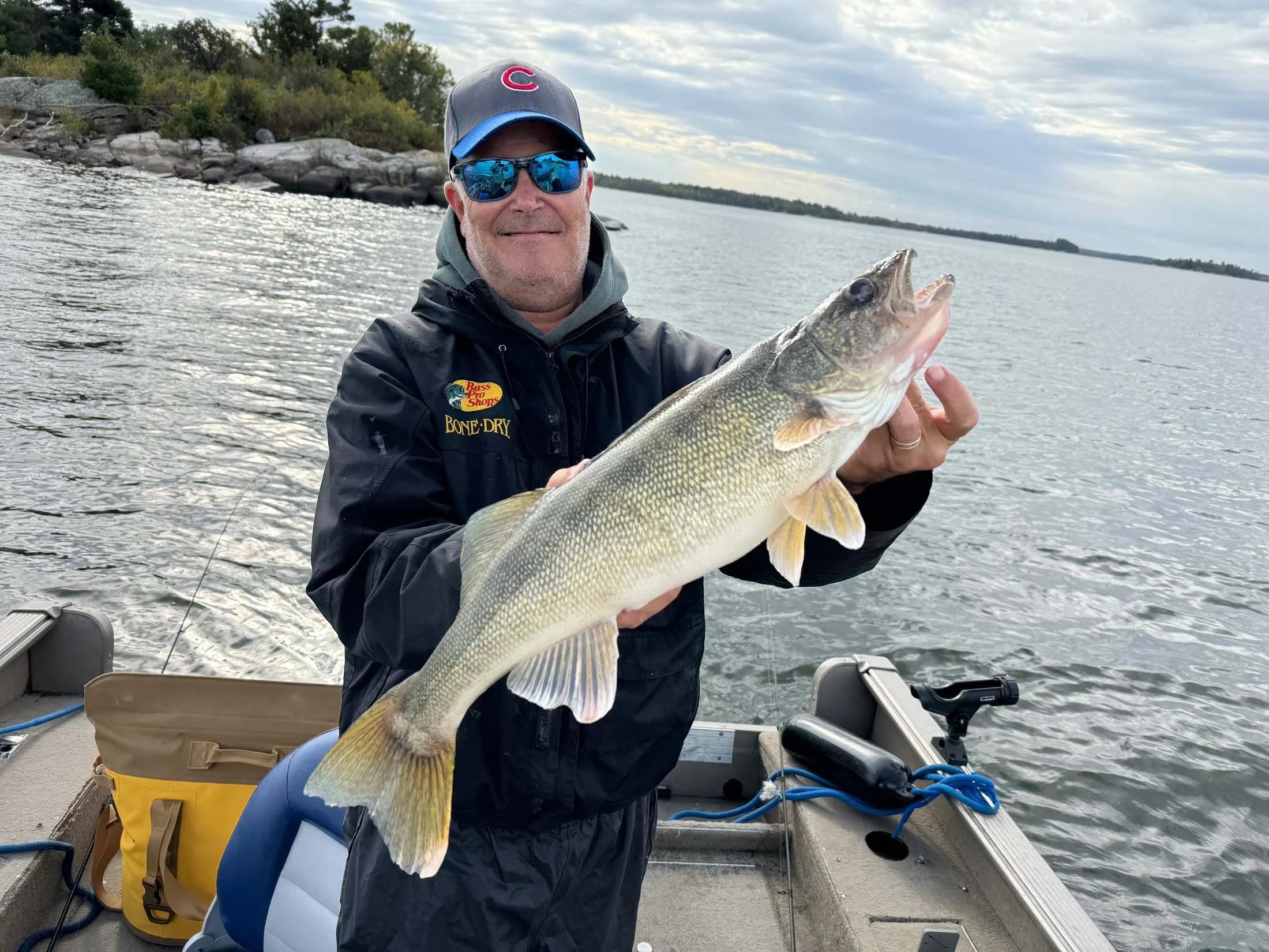 A man wearing sunglasses and a gray cap holding a large fish on a boat in a lake, with rocky shoreline and trees in the background.