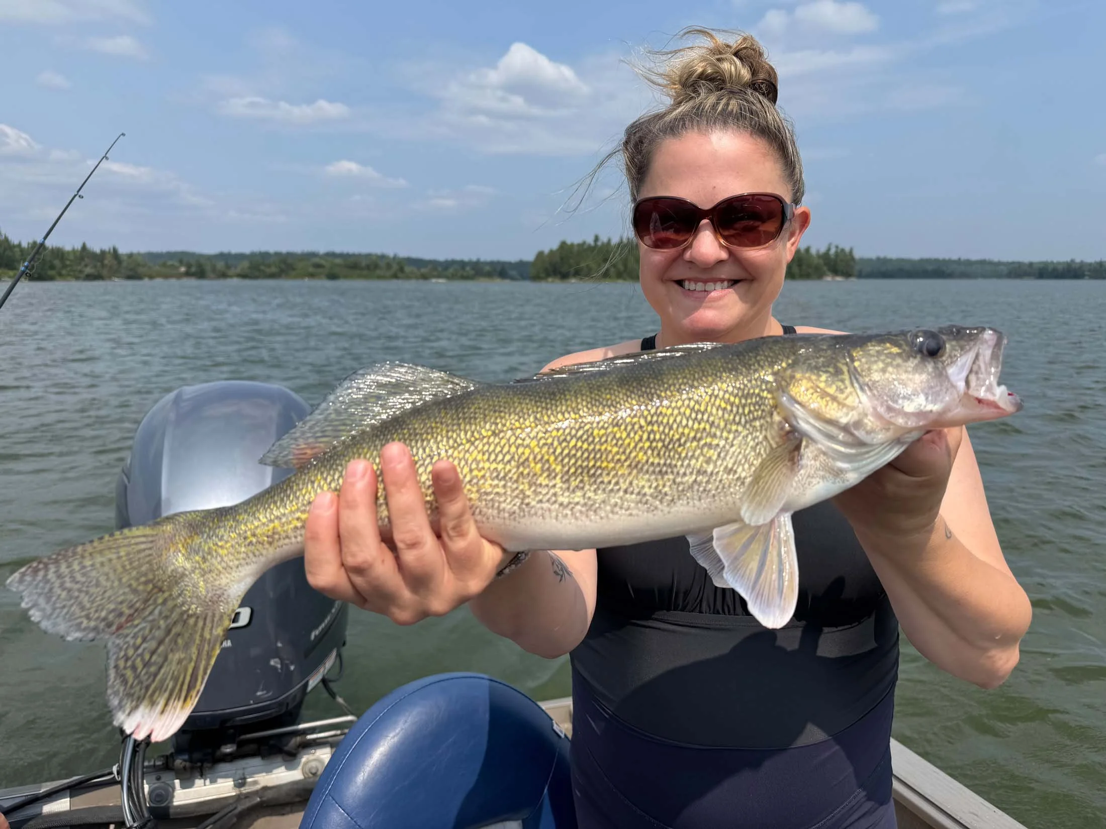 Woman in sunglasses smiling and holding a large fish on a boat with water and trees in the background.