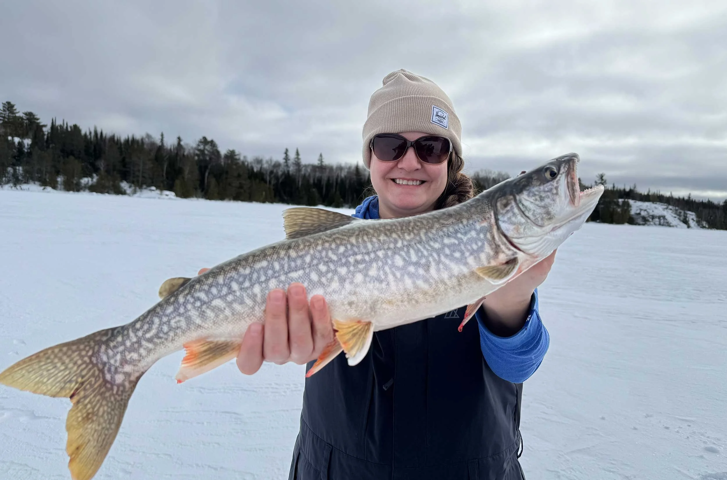 Person wearing sunglasses and a beige beanie holding a large fish over a snowy landscape with trees in the background.