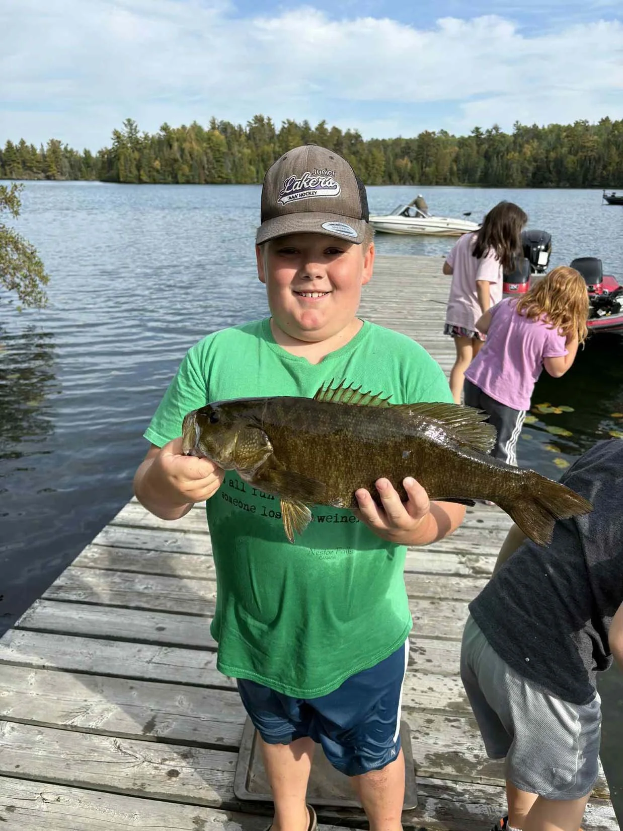 A young boy in a green shirt and a gray cap holding a large fish on a dock by a lake, with other children and boats in the background.