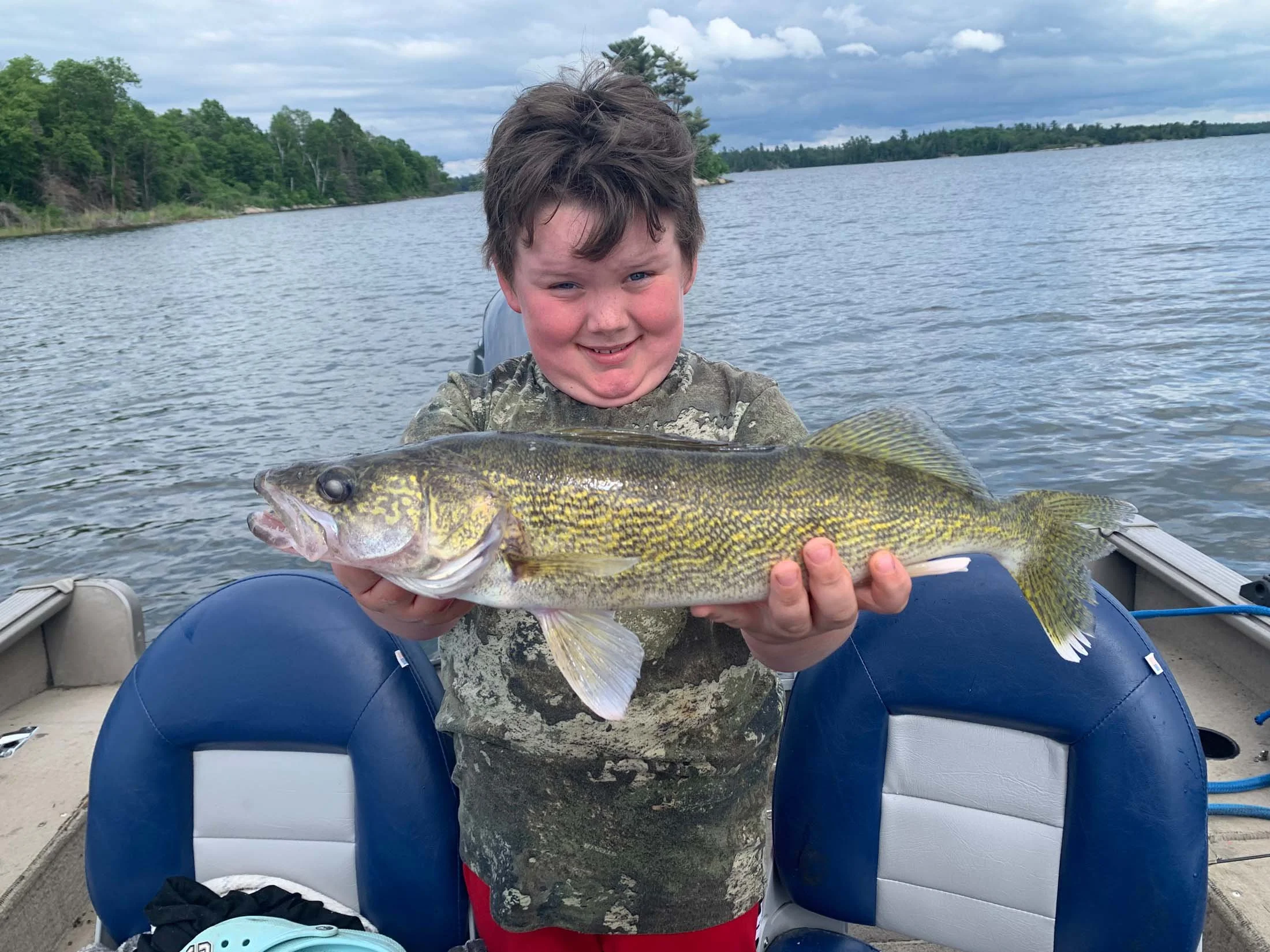 A young boy in a camouflage shirt is on a boat, holding a large fish he caught, with a body of water and green trees in the background.