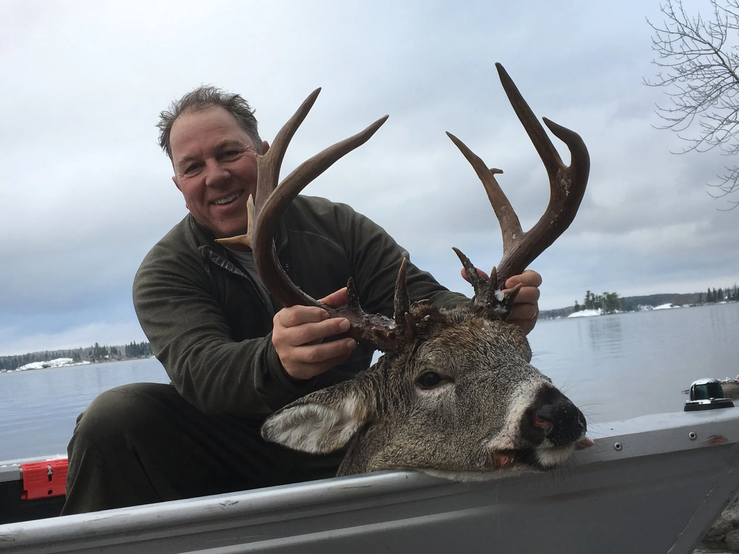 A man holding a large deer with antlers outdoors by a body of water on a cloudy day.