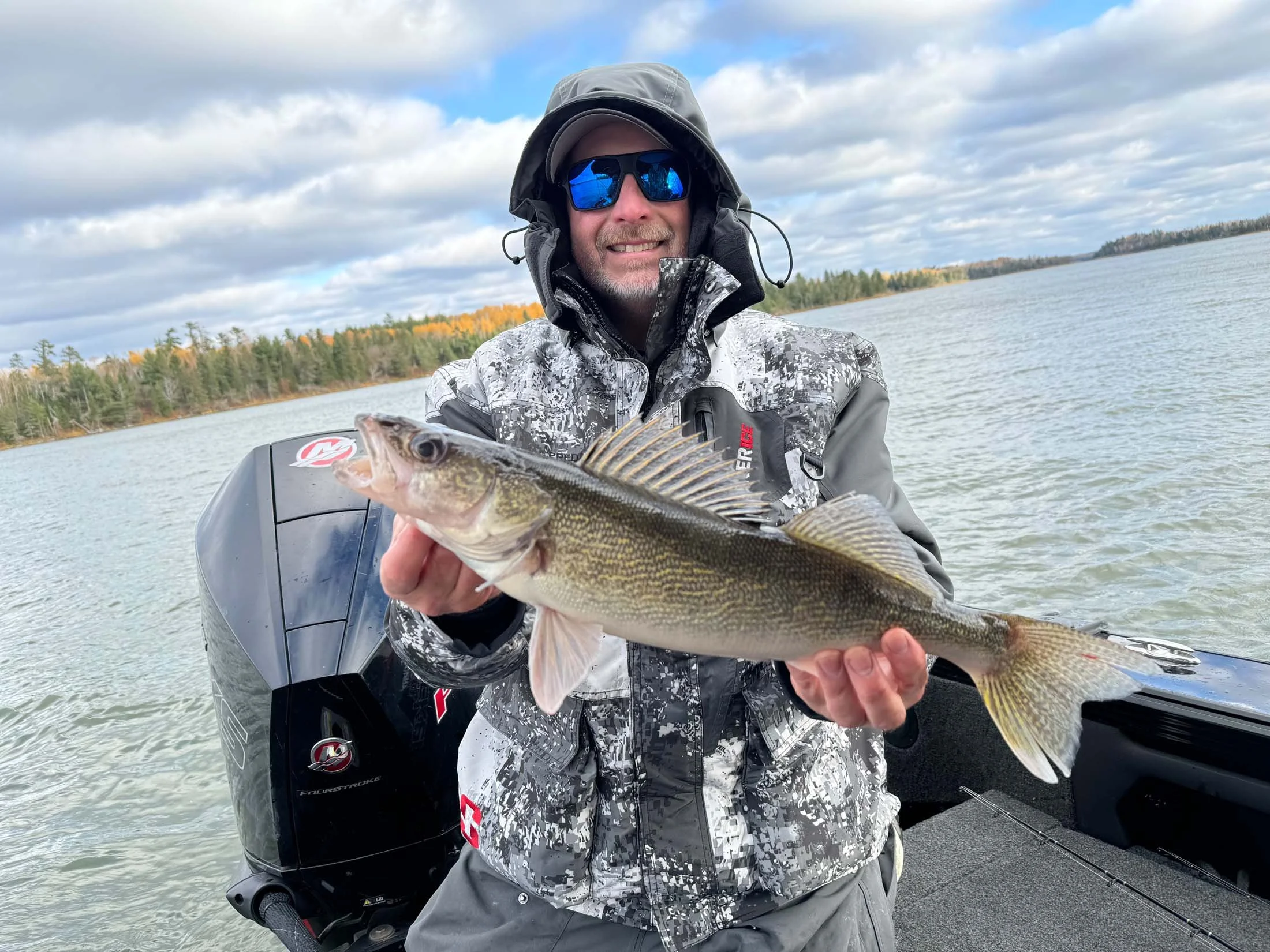 A man in a gray camouflage jacket, wearing sunglasses and a hood, is smiling and holding a large fish while sitting in a boat on a lake with a treeline and cloudy sky in the background.