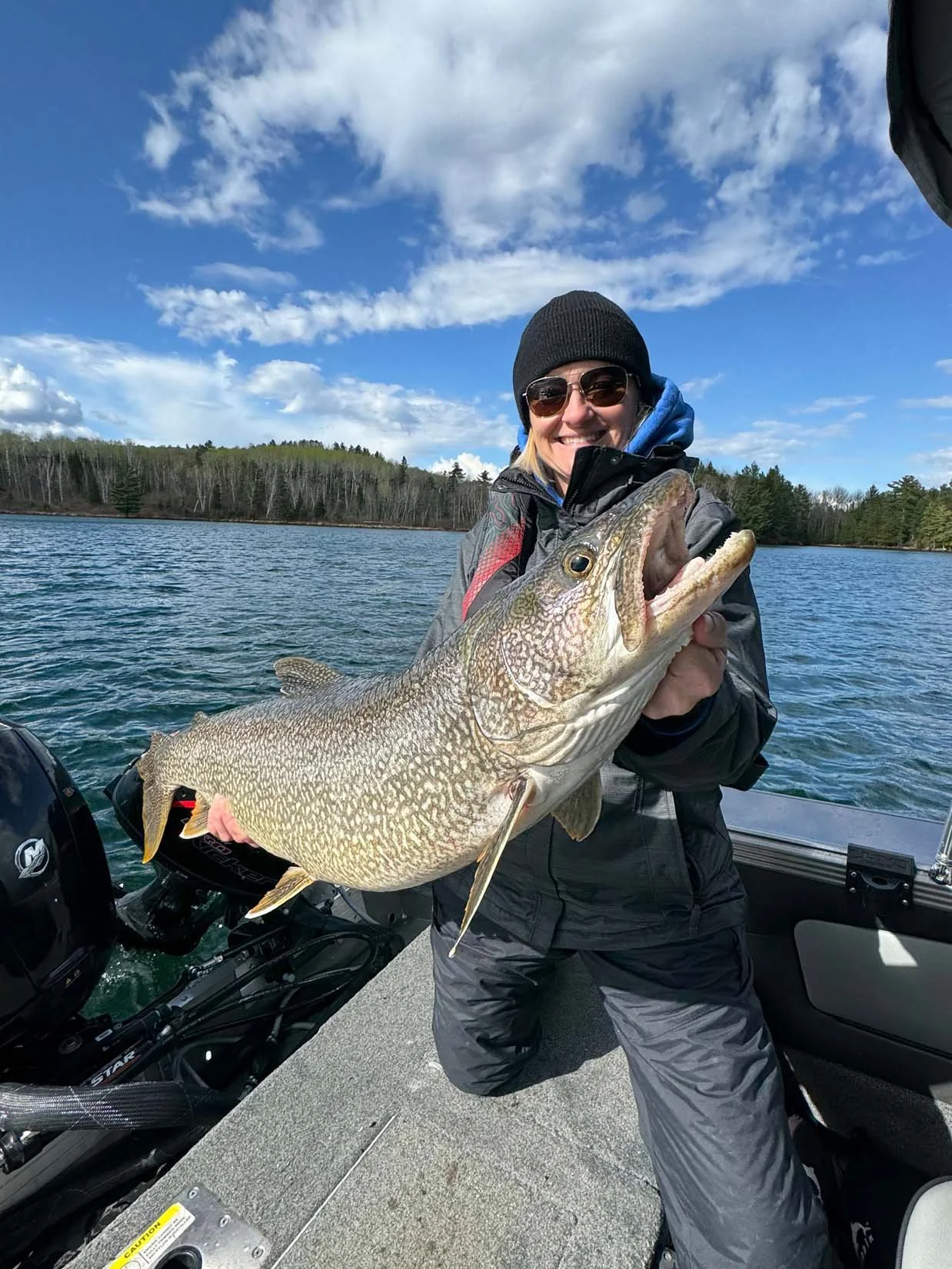 A woman with sunglasses and a beanie holding a large fish on a boat in a lake, with a backdrop of trees and a partly cloudy sky.