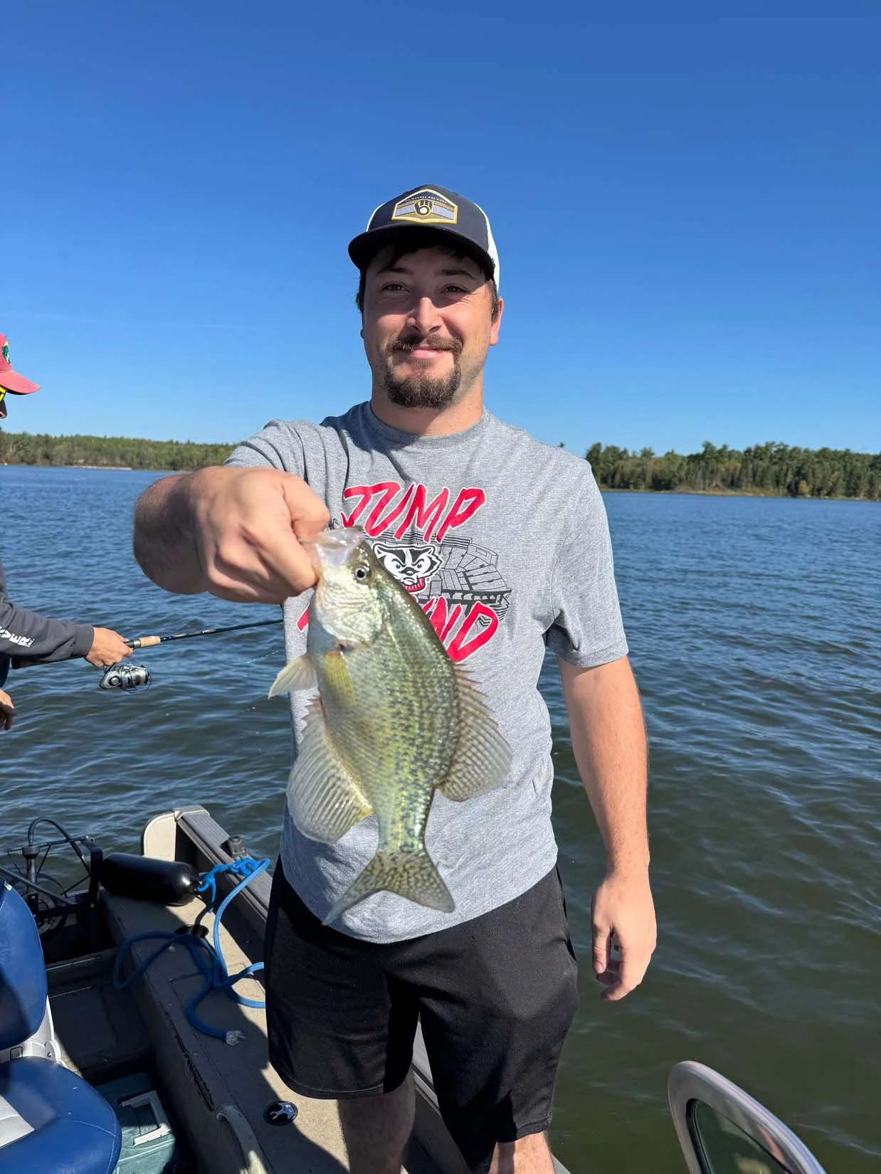 A man standing on a boat by a lake, holding up a large fish he caught. He is smiling and wearing a gray T-shirt with red lettering and a black and white baseball cap. In the background, another person is fishing on the lake.