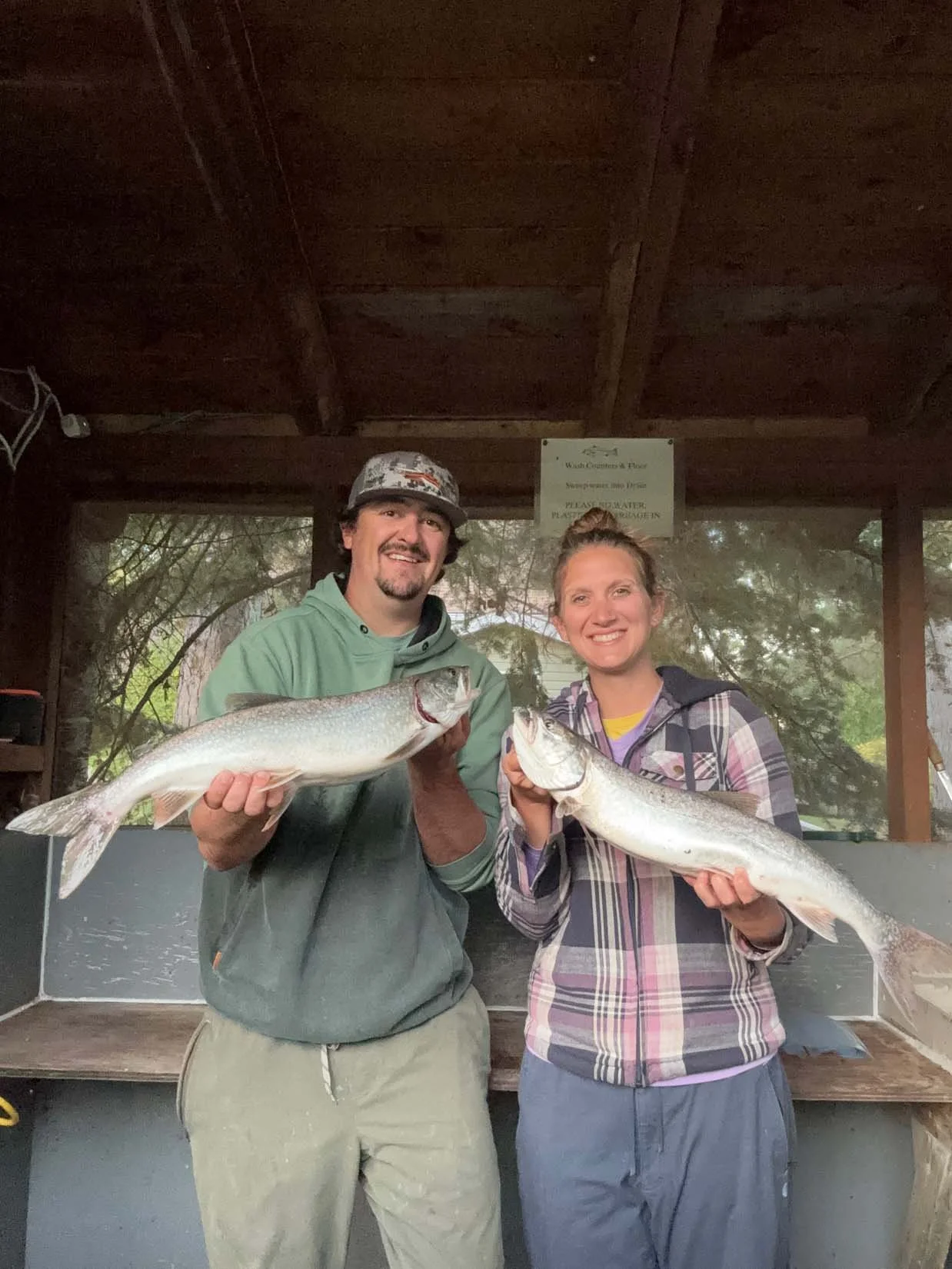 A man and a woman smiling and holding two large fish they caught, standing inside a rustic outdoor shelter with trees visible in the background.
