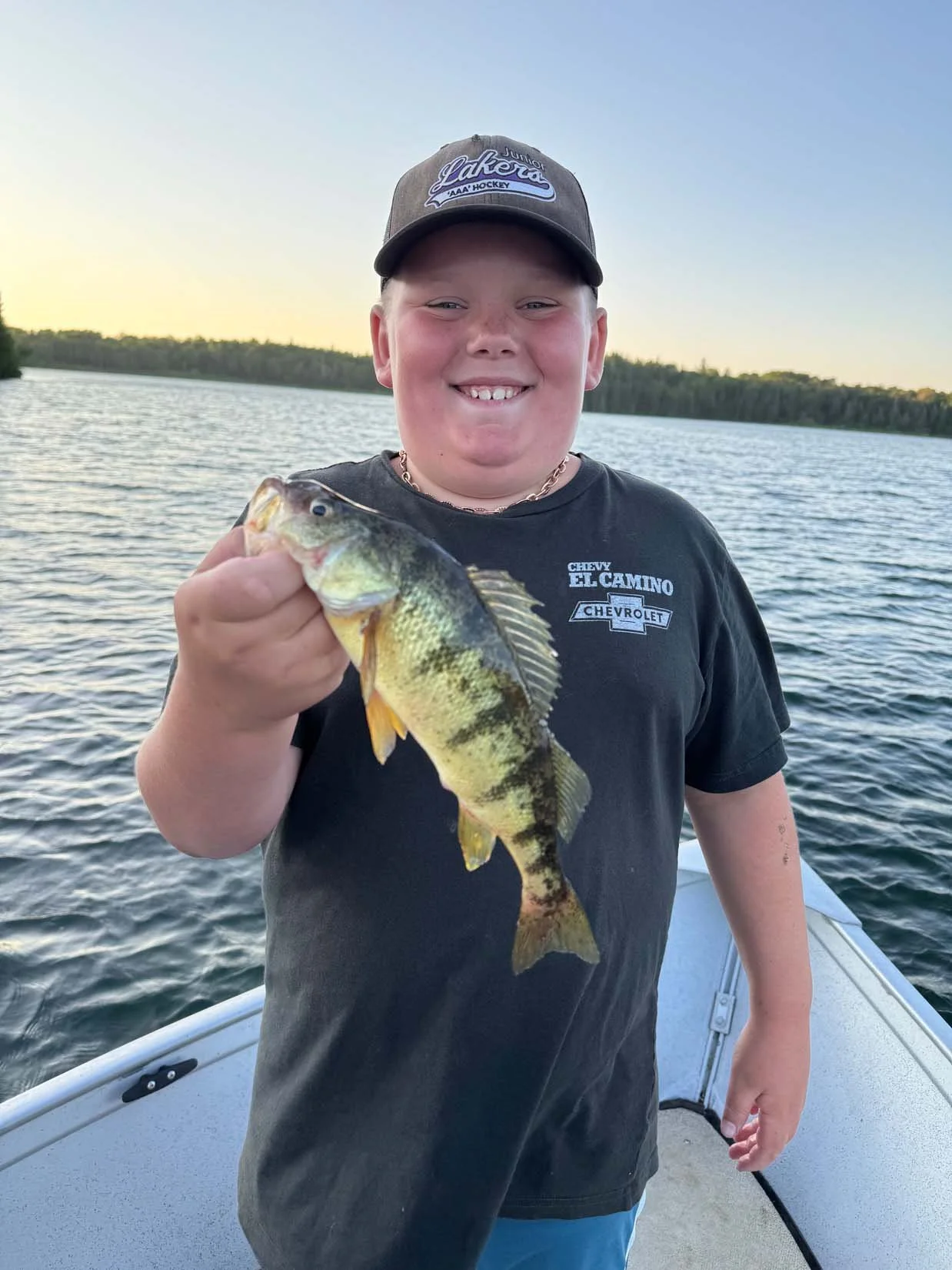 A young boy smiling and holding a largemouth bass fish on a boat, with a lake and forested shoreline in the background during sunset.