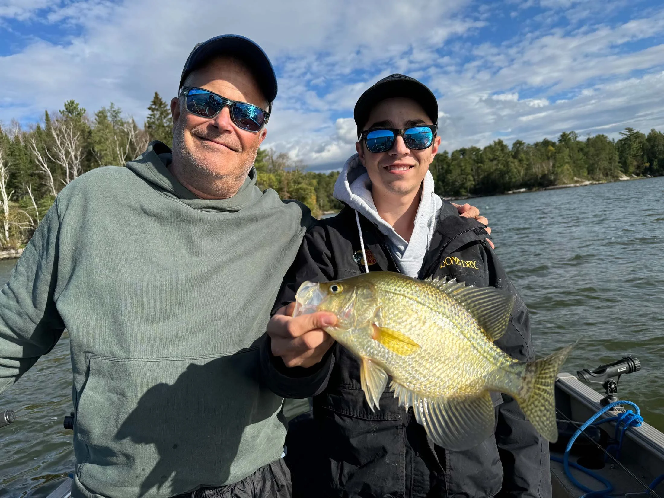 Two men wearing sunglasses and caps on a boat by a lake, holding a fish, with trees and a partly cloudy sky in the background.