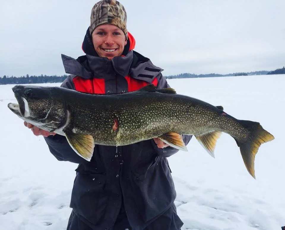 A smiling man dressed in winter outdoor gear holding a large fish on a snowy landscape.