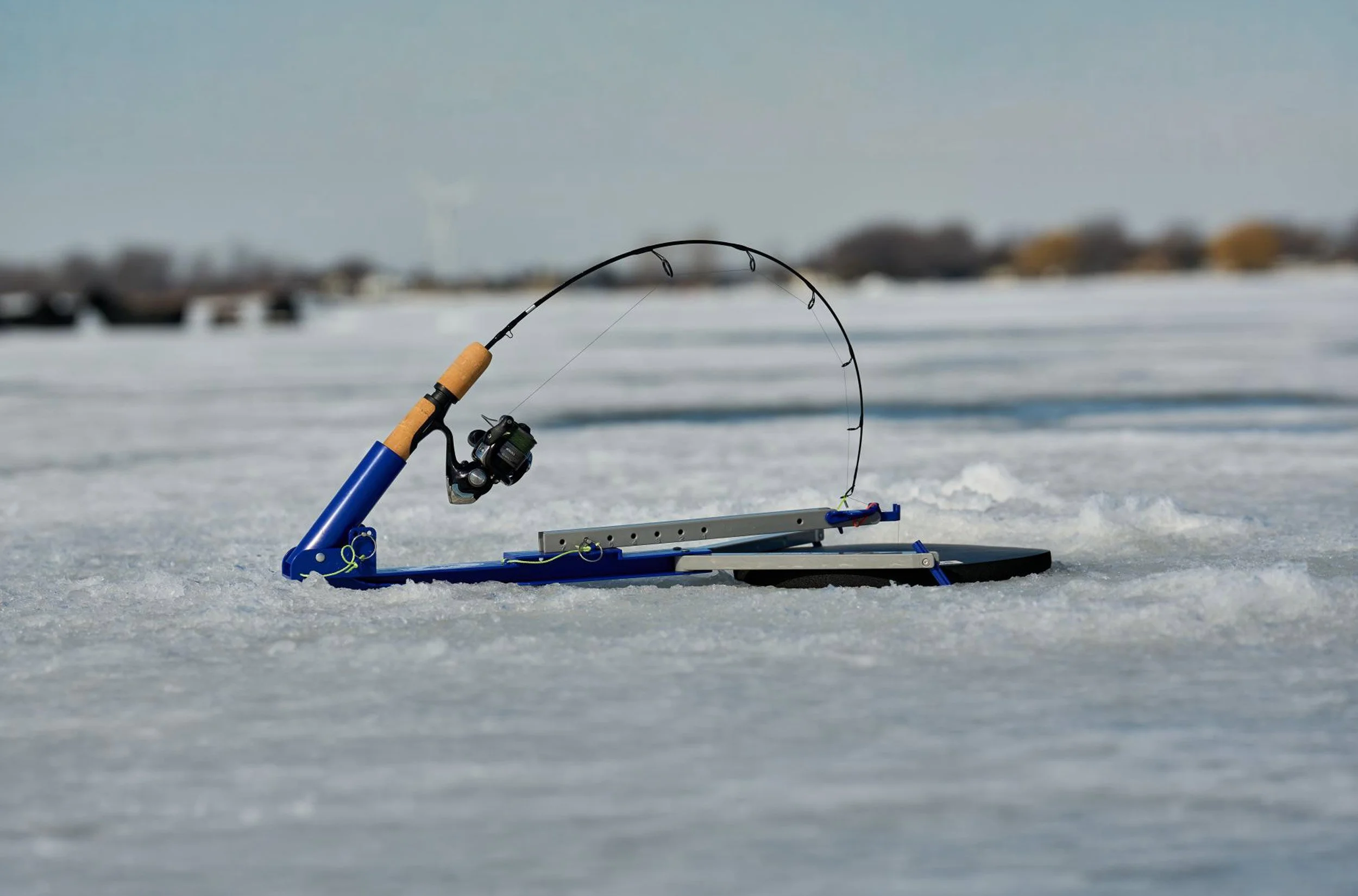 A fishing rod with a reel is set up on a small blue fishing kayak on snowy ice, against a blurred background of distant buildings and a wind turbine.