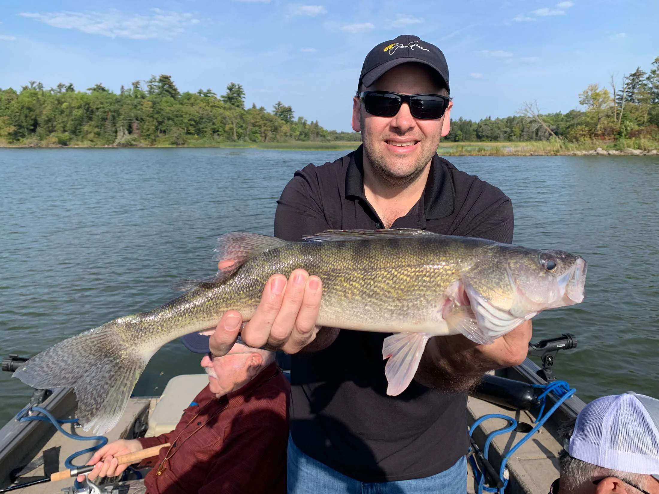 A man in sunglasses and a black cap holding a large fish, with a person in the background on a boat on a lake surrounded by trees.