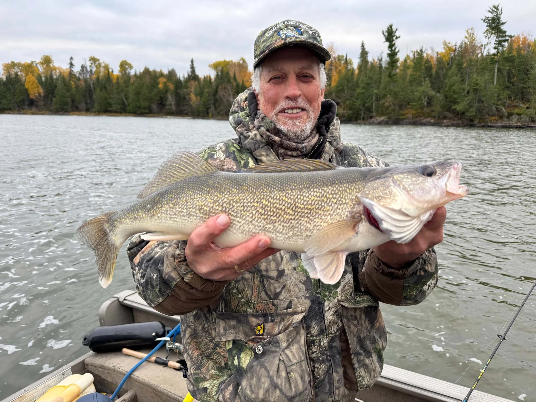 A man in camouflage clothing and a cap is holding a large fish on a boat near a lake with trees in the background.