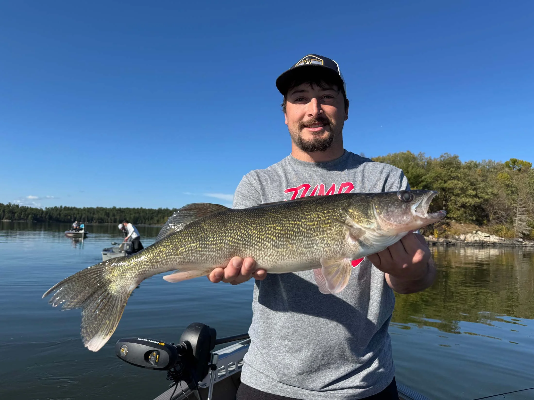 A man with a beard and mustache wearing a gray t-shirt and a cap, holding up a large fish with a dark green and yellowish pattern, standing near a body of water with trees in the background and a few people fishing in the distance.