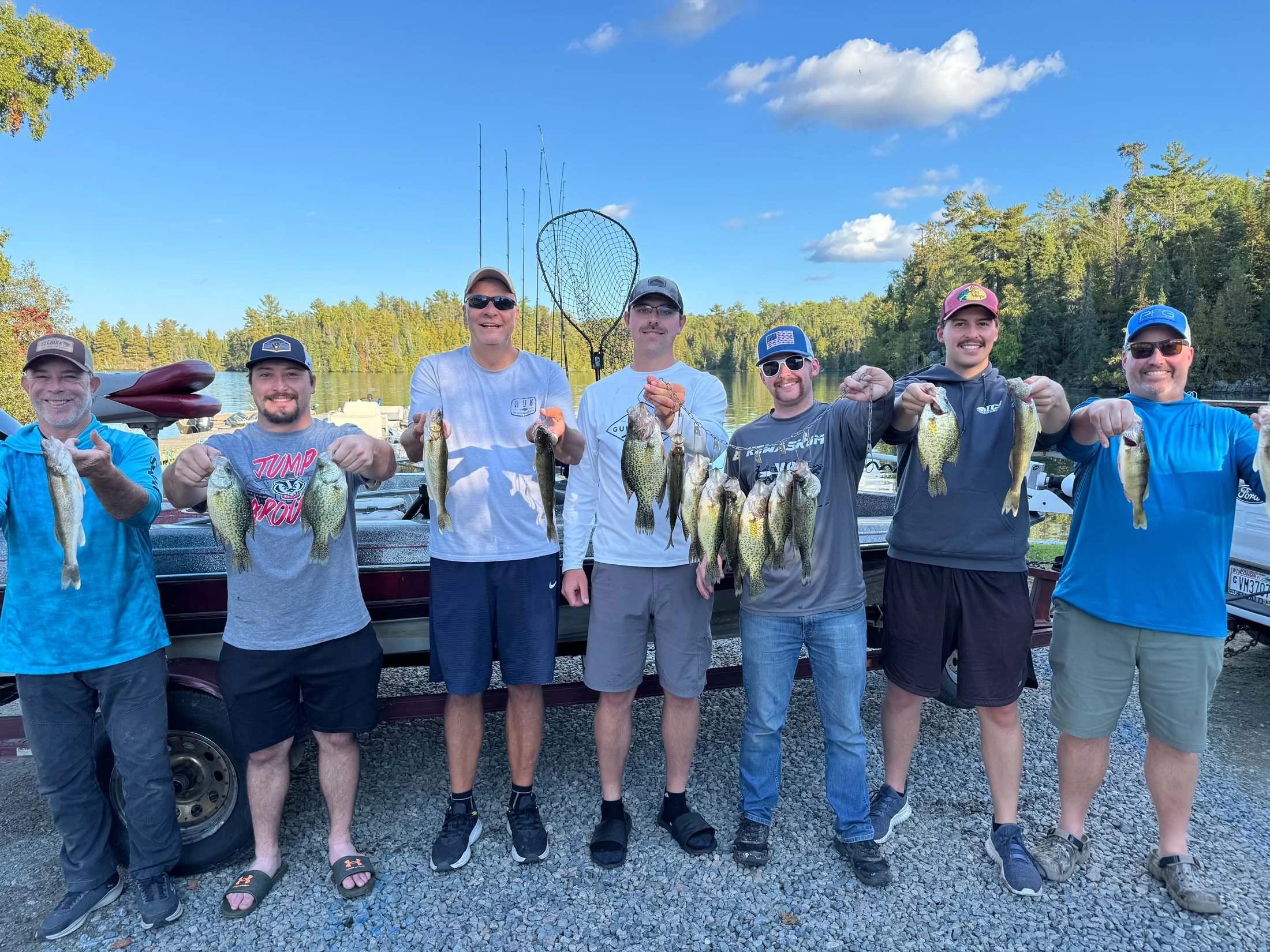 Group of seven men standing outdoors near a lake, holding up fish they caught during a fishing trip, with trees and blue sky in the background.
