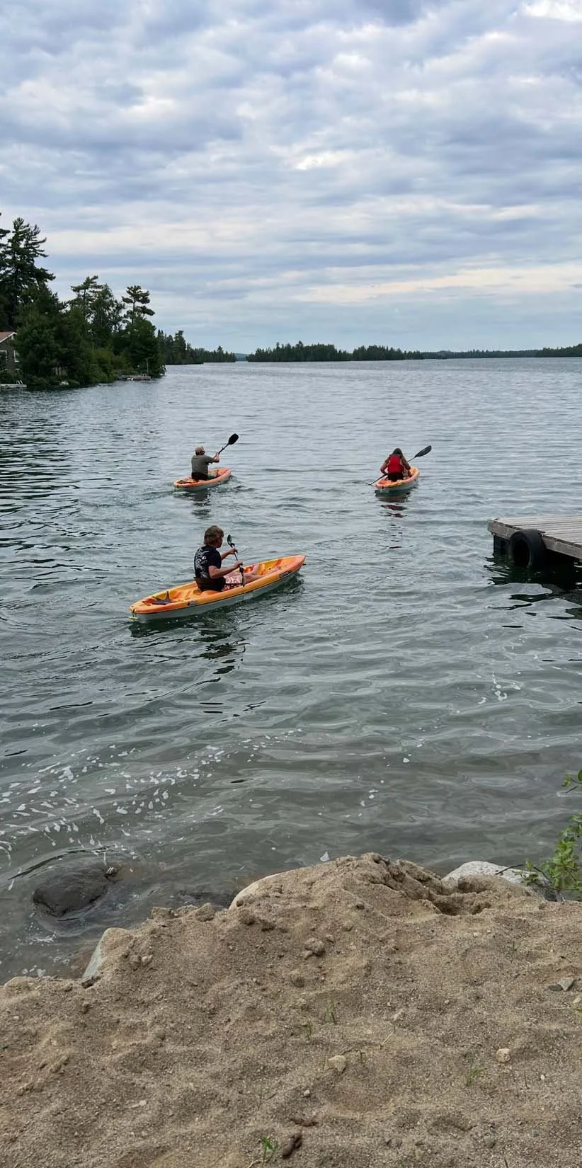 Three people kayaking on a calm lake, with a sandy shoreline in the foreground and a cloudy sky above.