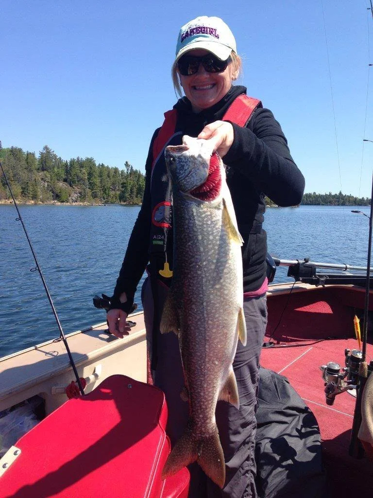 A woman on a boat holding a large fish she caught while fishing on a lake, smiling under a white cap and sunglasses.