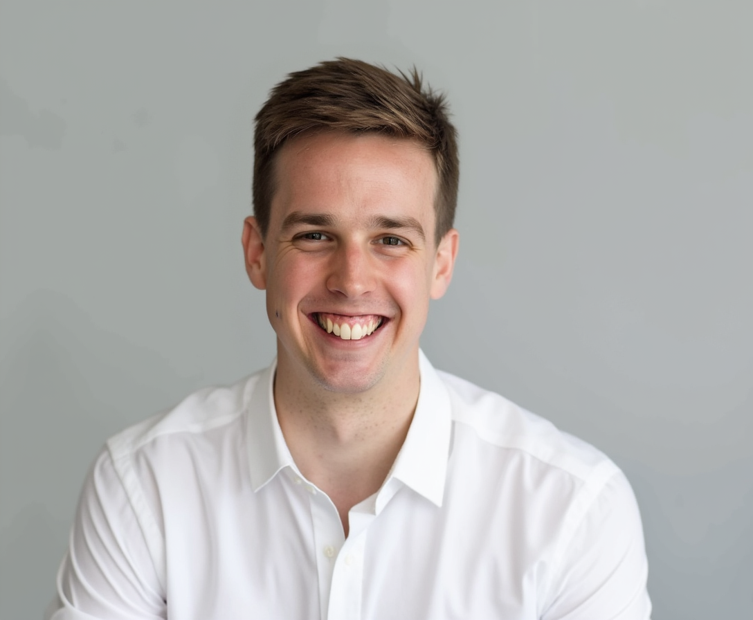 A young man with brown hair wears a white shirt and smiles in front of a plain gray background.