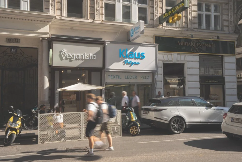 Straßenszene mit Geschäften, darunter Veganista und Klaus Pfeifer, sowie Autos und Fußgänger in einer Stadt, vermutlich Wien.