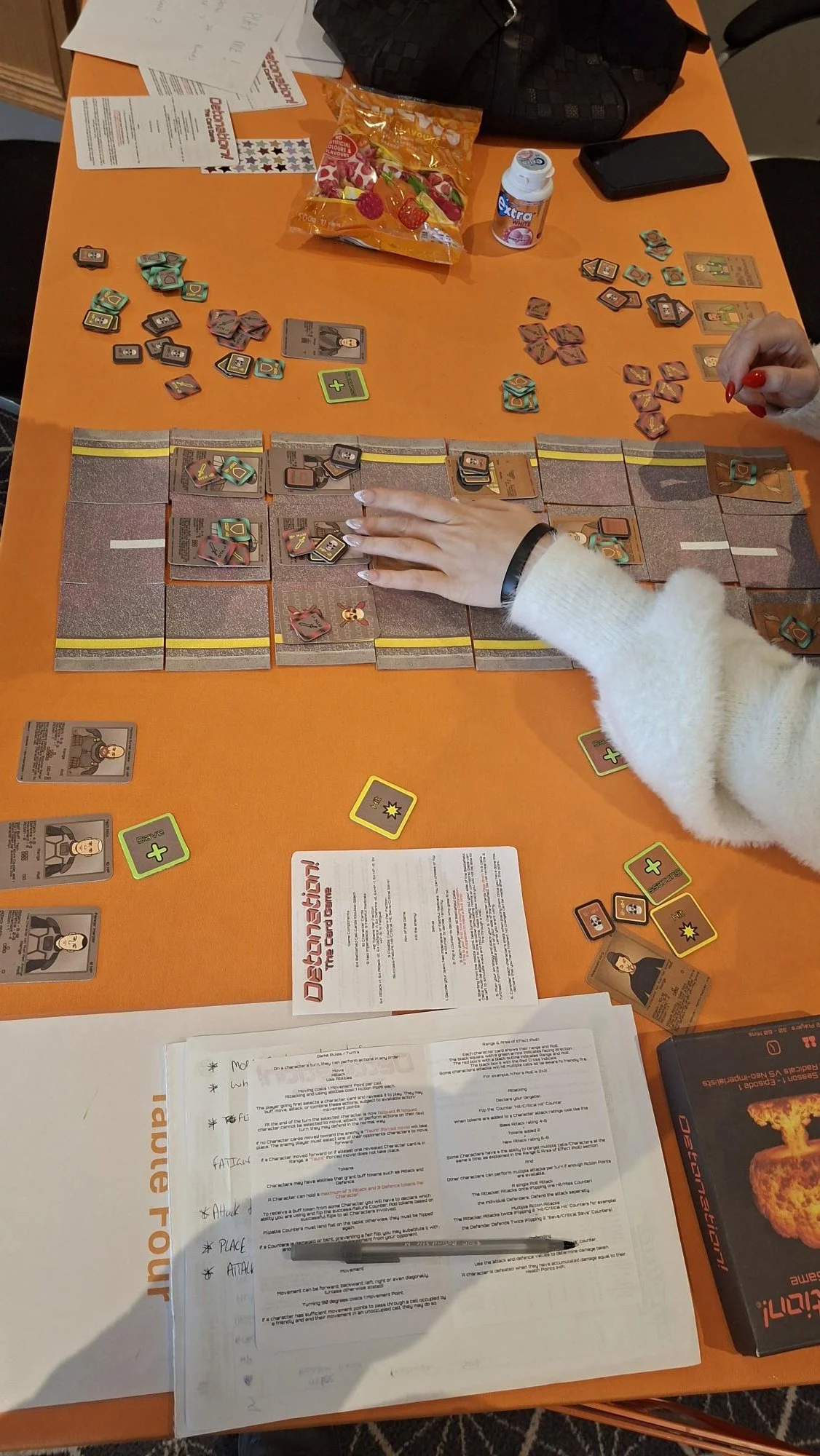 People playing the card game 'Detonation! The Card Game' at Handycon Jan 2026. a table with game pieces, cards, and instruction sheets.