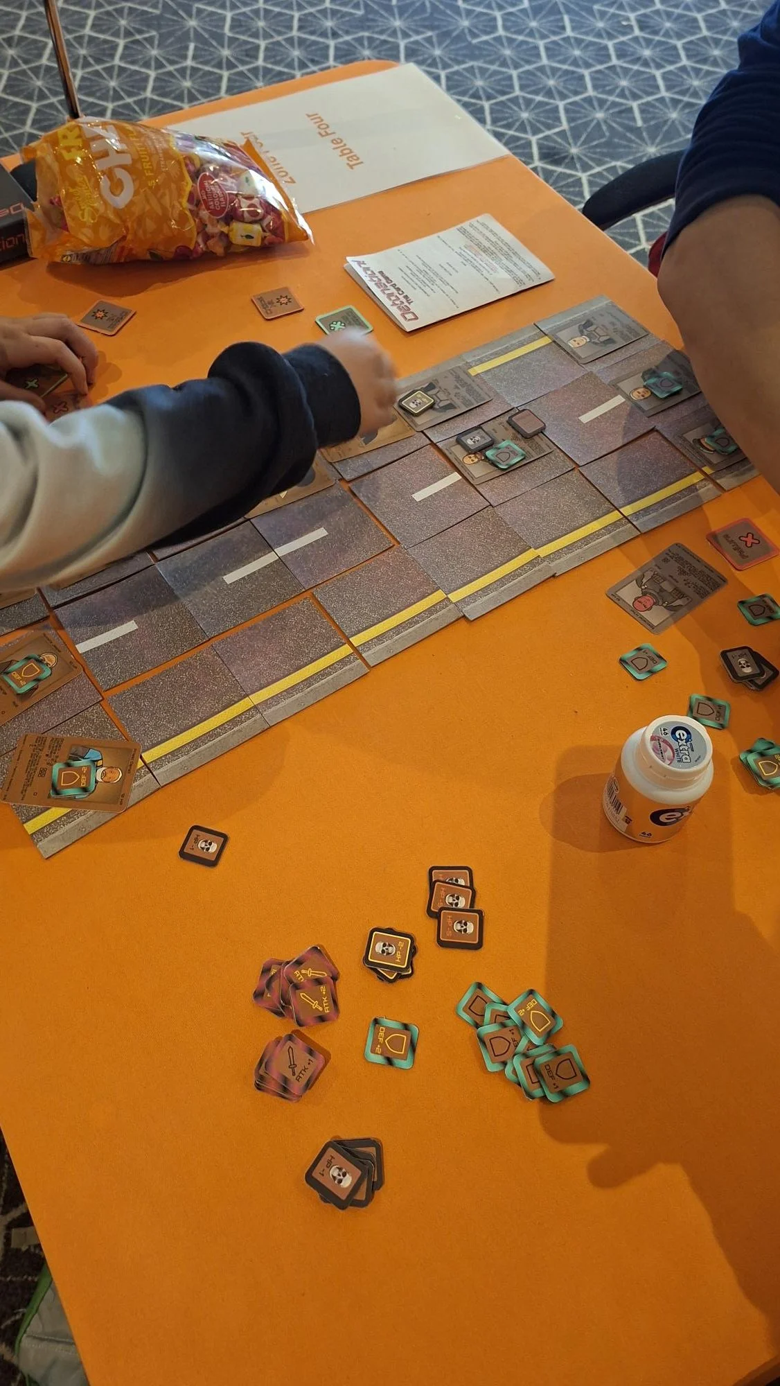 People playing the card game 'Detonation! The Card Game' at Handycon Jan 2026. a table with game pieces, cards, and instruction sheets.