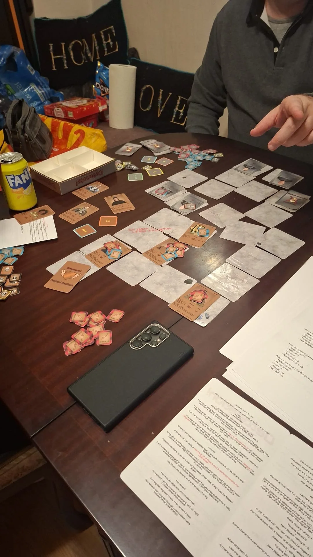 People playing the card game 'Detonation! The Card Game' December 2025. a table with game pieces, cards, and instruction sheets.