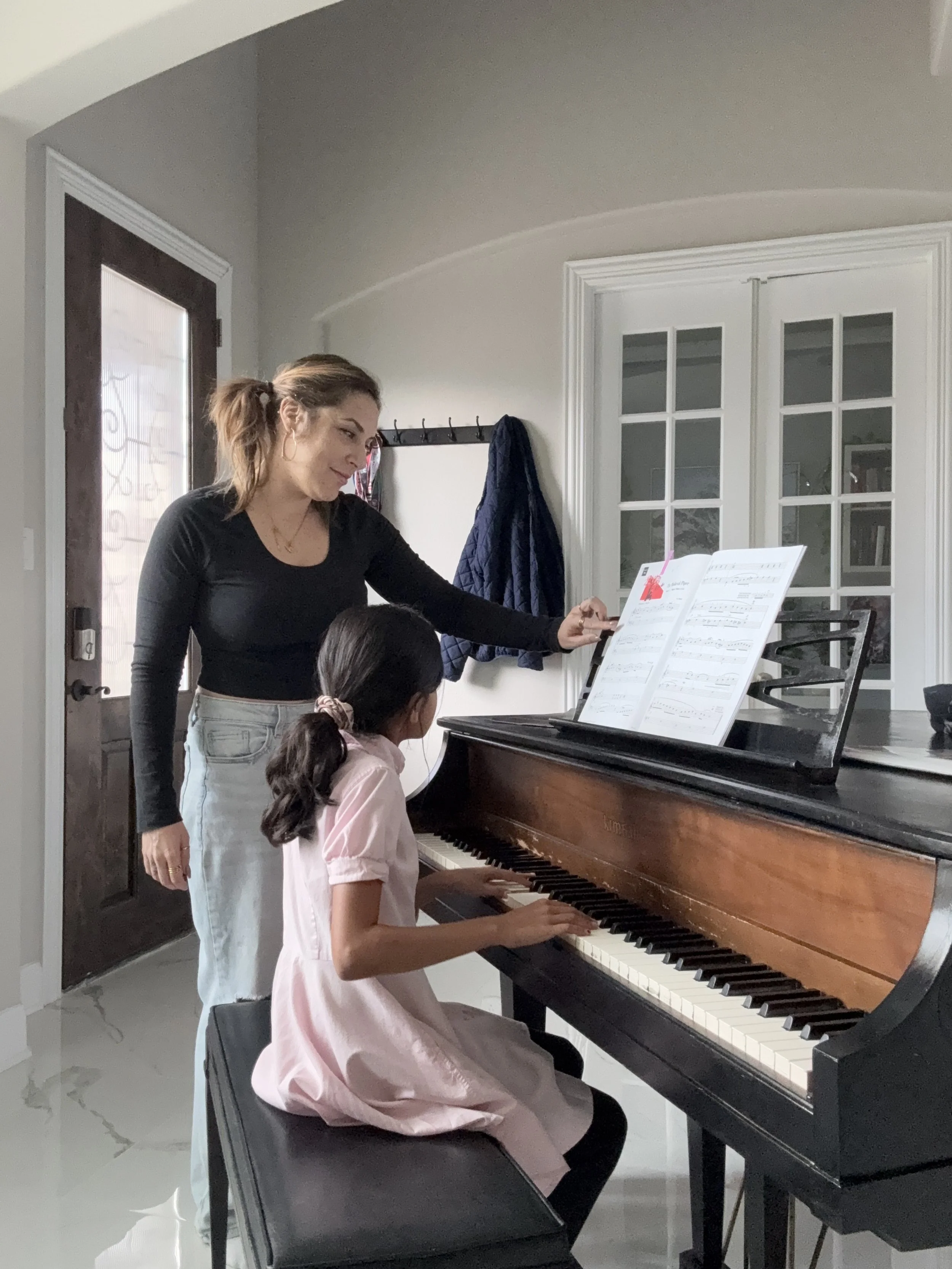 A woman teaching a young girl how to play the piano in a brightly lit room, with sheet music on the piano.