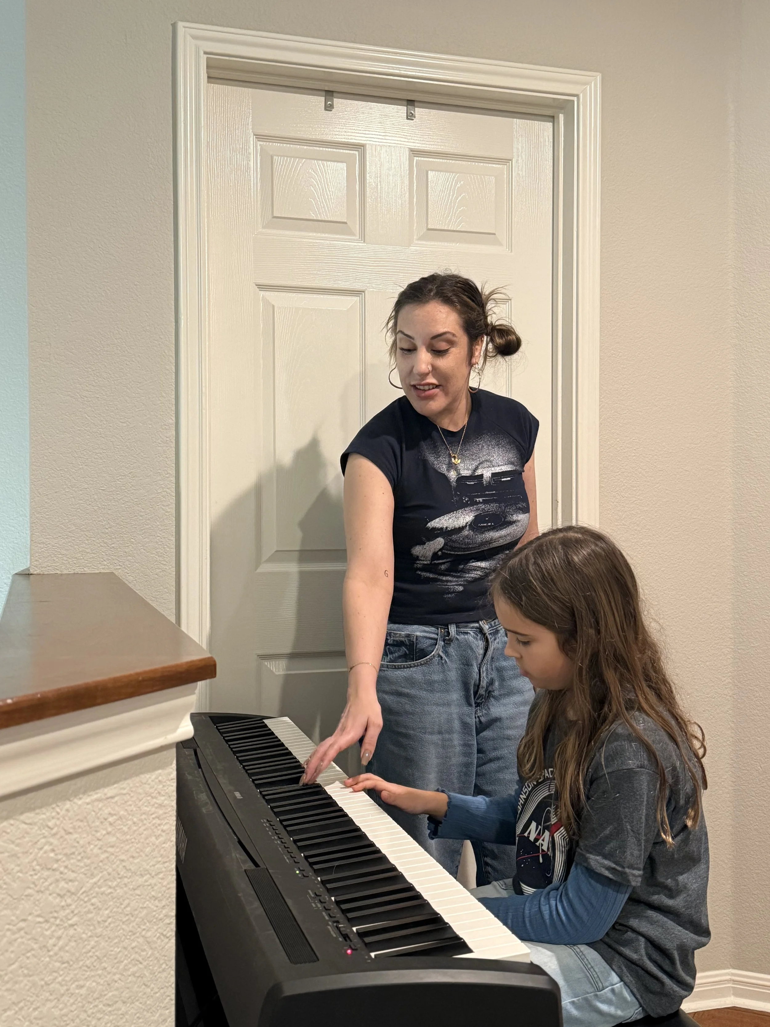 A woman teaching a young girl how to play the piano in a room with beige walls and a white door.