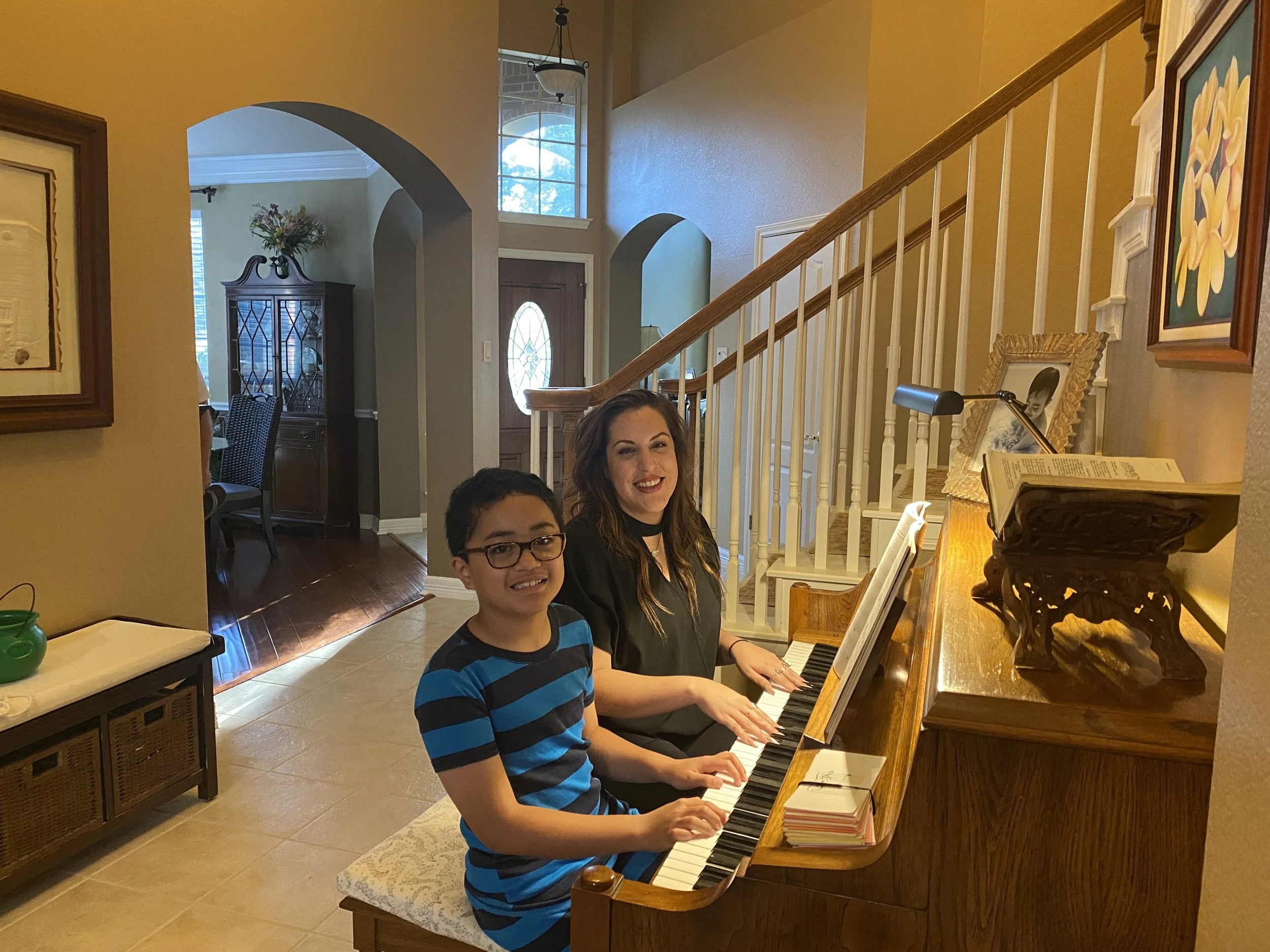 A woman and a young boy playing piano together in a cozy home interior, smiling at the camera.