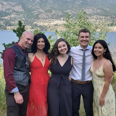 Five people standing outdoors near a lake with mountains and trees in the background, smiling and dressed in semi-formal attire.