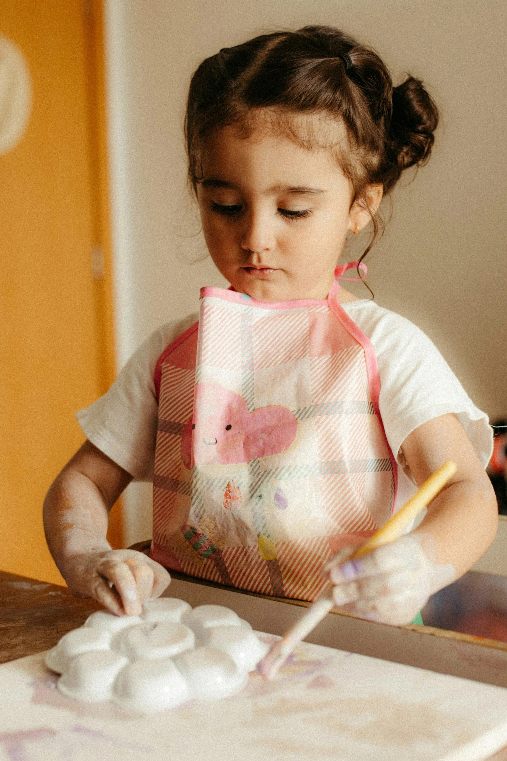 A young girl with dark hair styled in buns wearing a pink checkered apron with a bunny design, concentrating while making crafts with white foam balls at a table.
