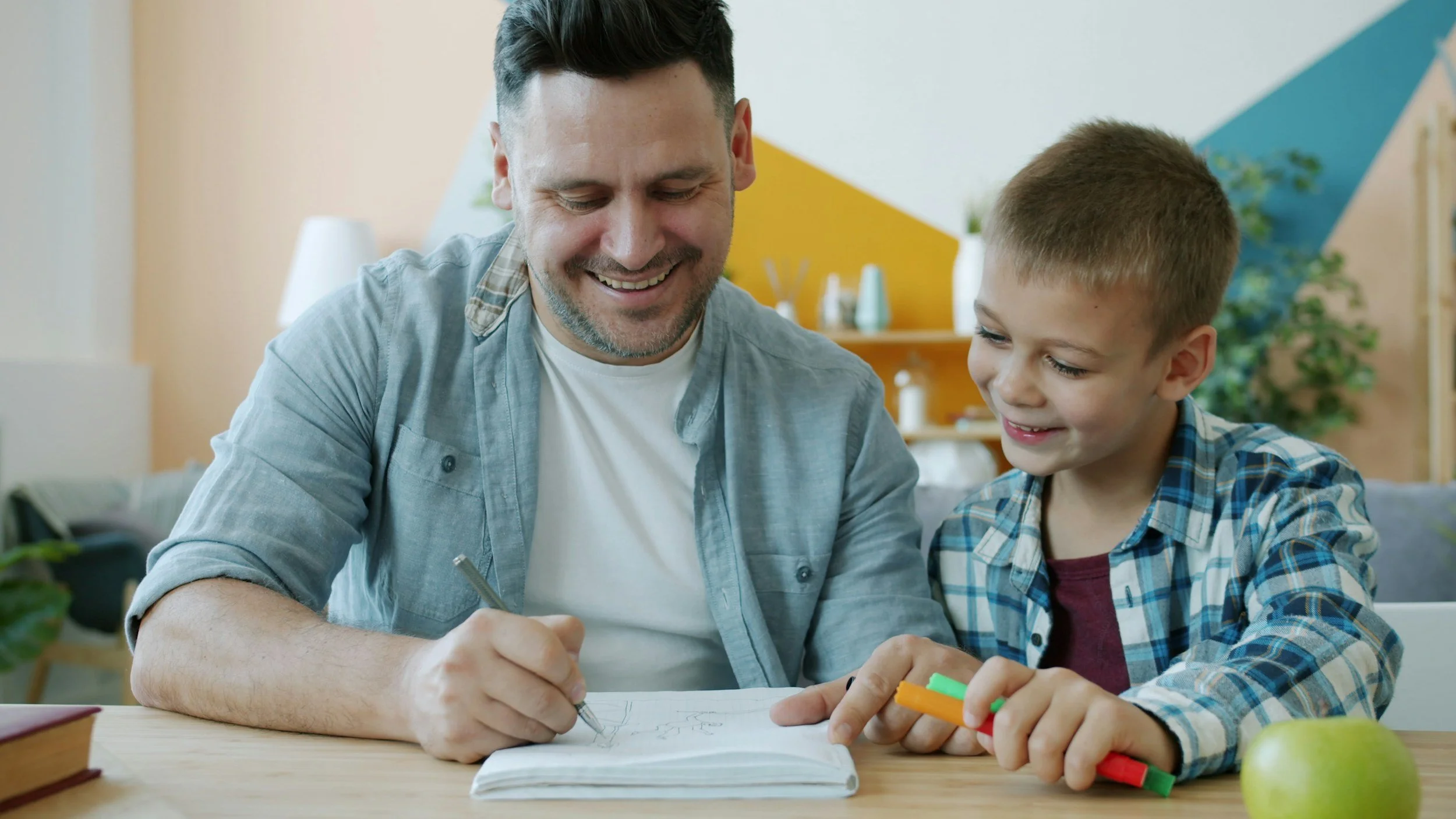 A man and a young boy sitting at a table, smiling and drawing together with colored markers.