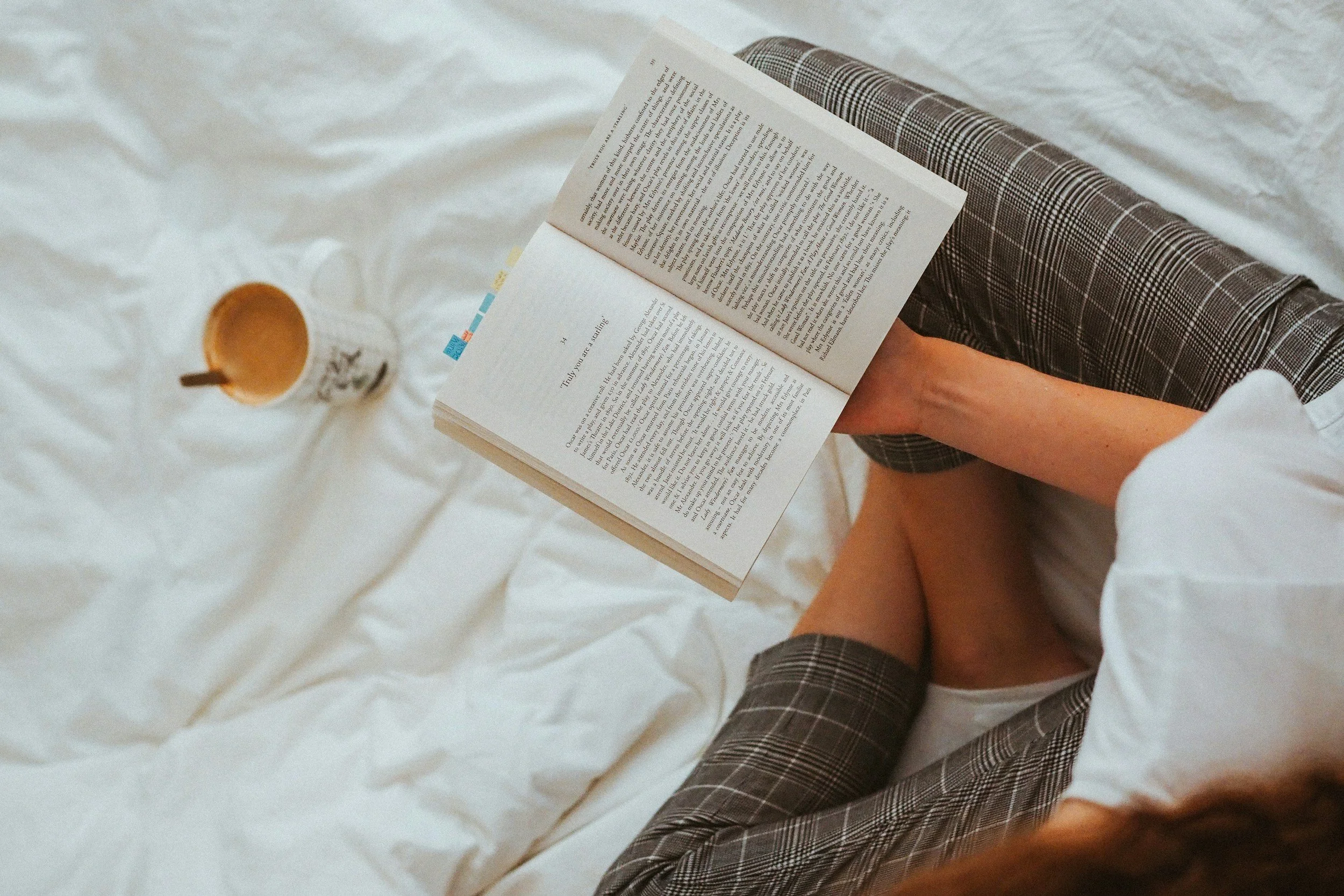 A person reading a book while sitting on a bed with white sheets, with a cup of coffee or tea nearby.