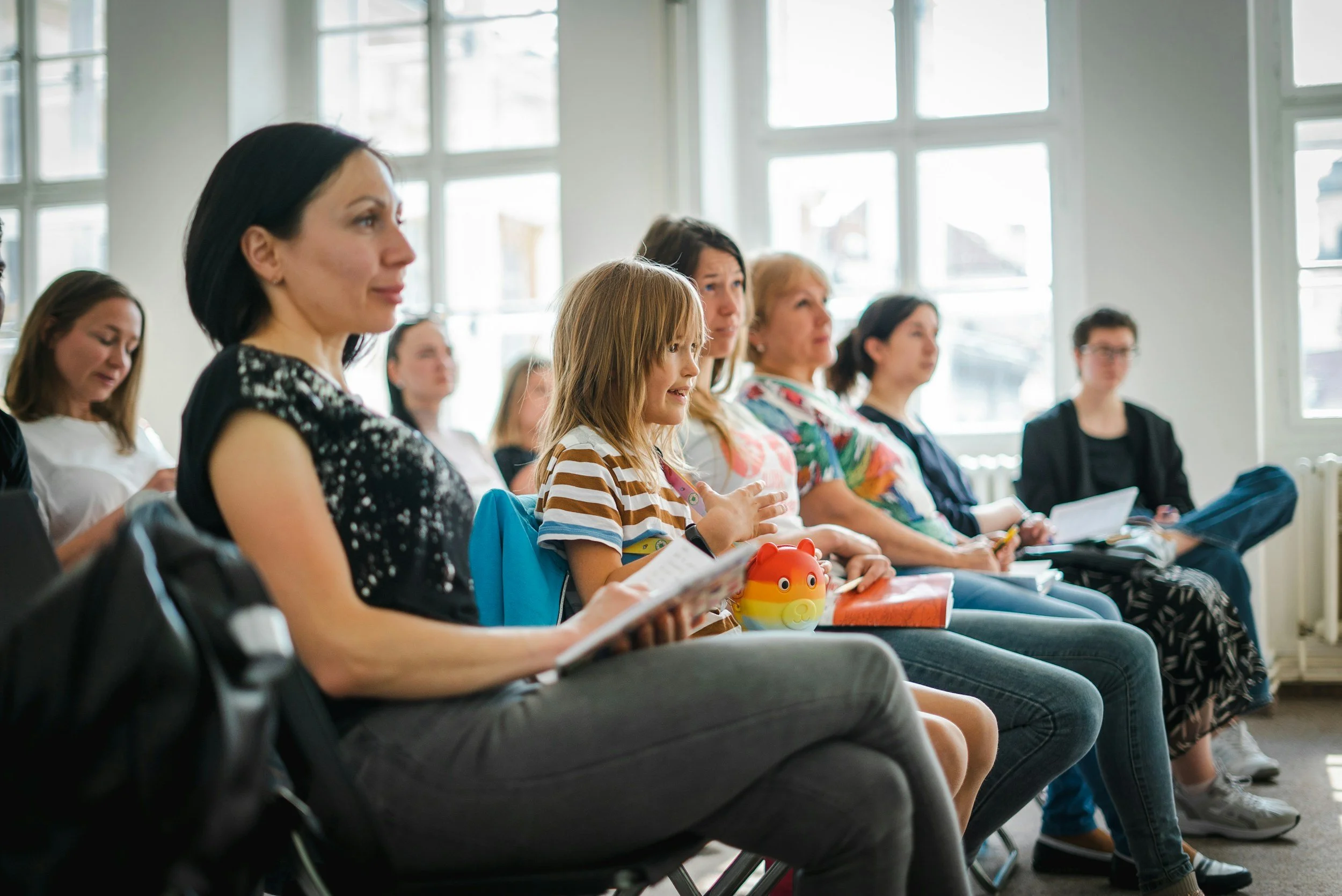 People sitting in chairs attentively listening in a bright room with large windows.