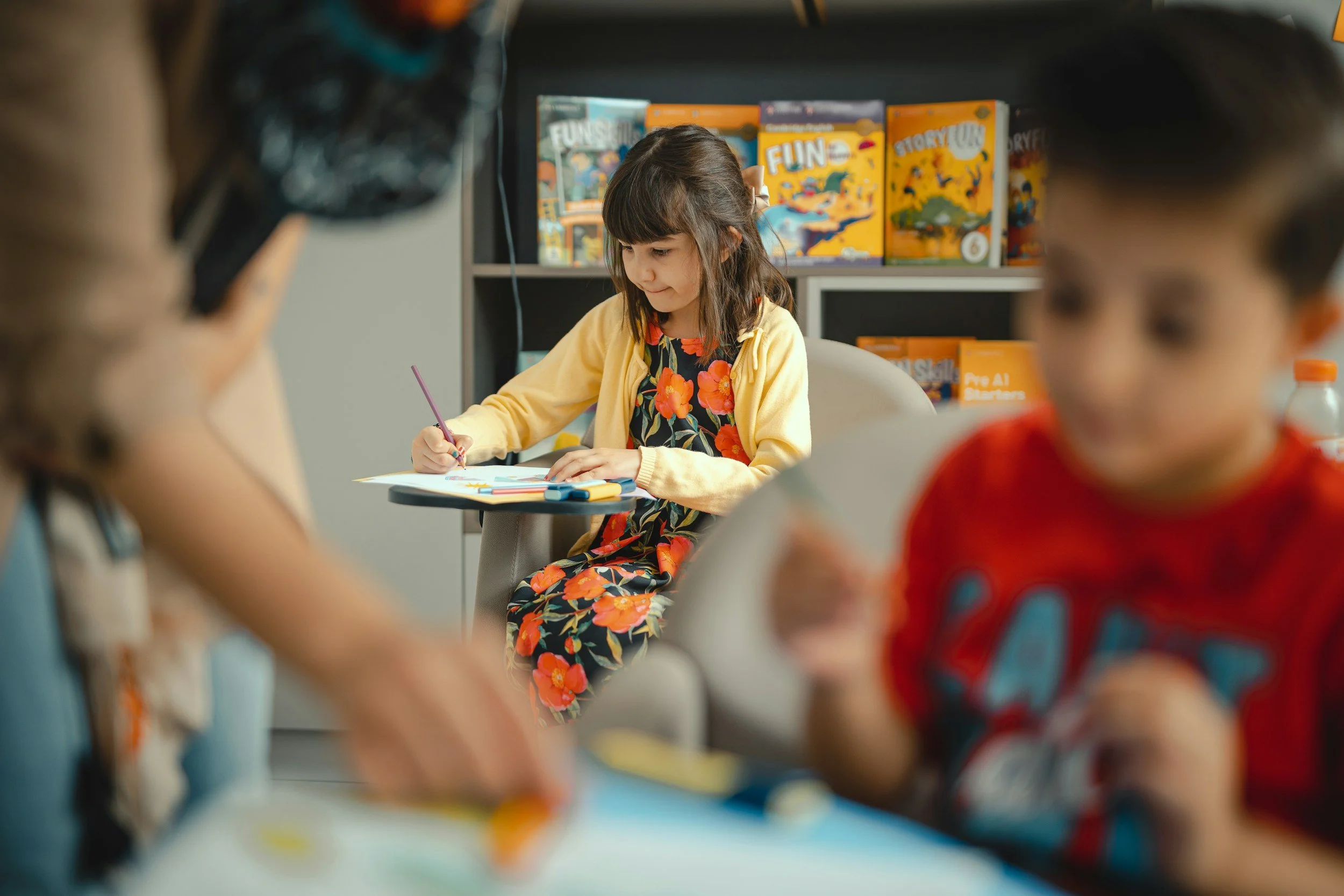 Young girl in floral dress and yellow cardigan sitting at a table writing with a pink pencil in a classroom or library setting, with bookshelves in the background, partially obscured by other people in the foreground.