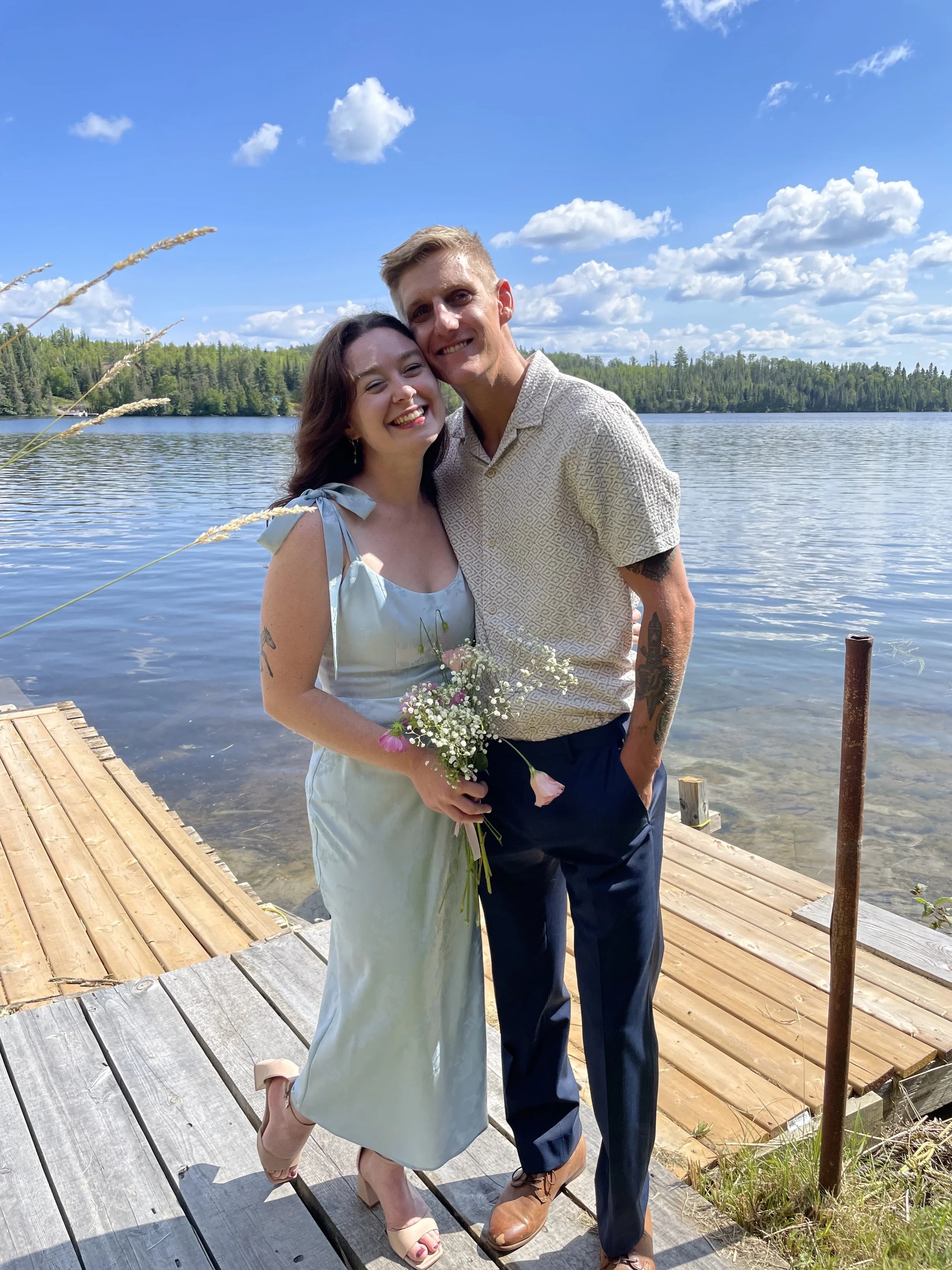 A smiling couple enjoying their time outdoors by a lake under a bright blue sky with scattered clouds. The woman is wearing a light dress with tie shoulder straps and holding a small bouquet of flowers, while the man is dressed in a patterned short-sleeve shirt and dark pants. They are standing on a wooden dock with a wooded shoreline in the background.
