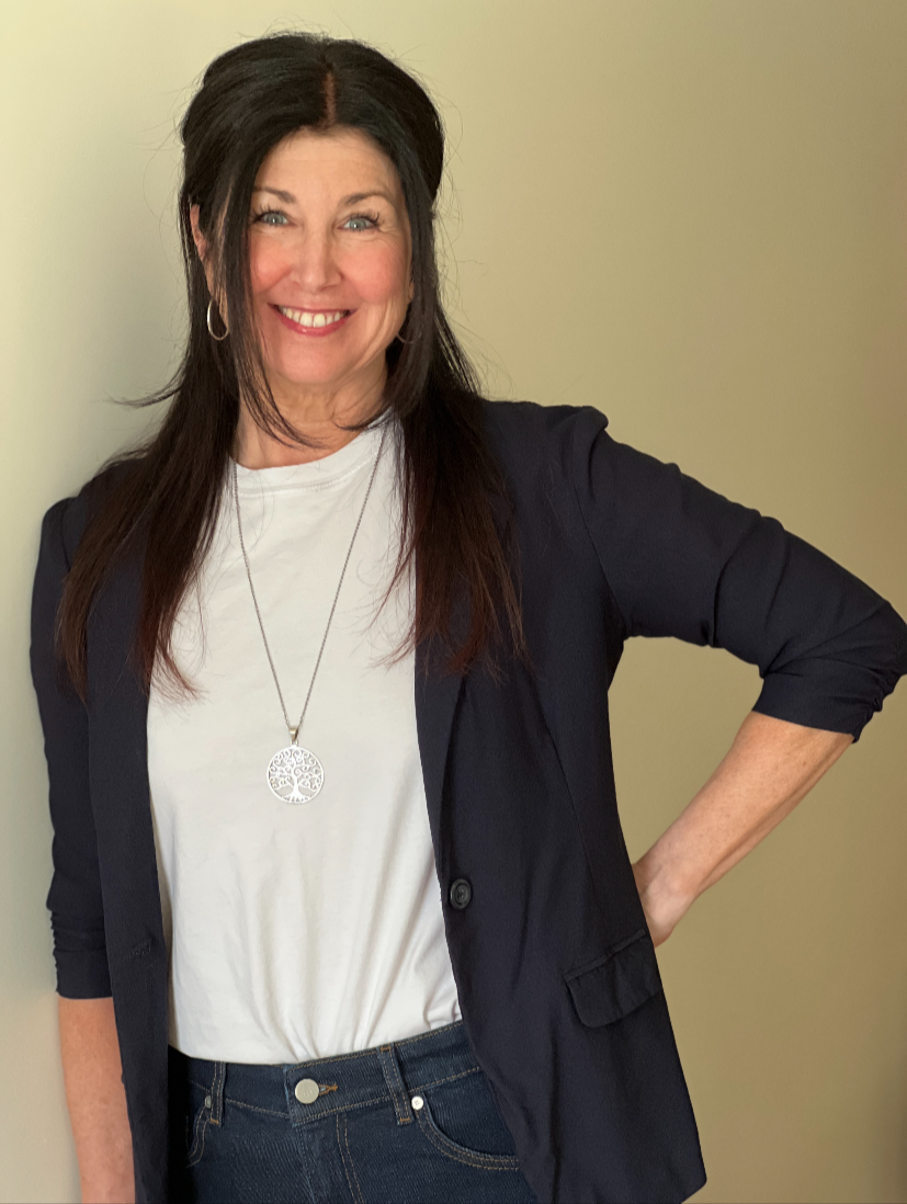 A woman with long dark hair, wearing a black blazer over a white t-shirt, blue jeans, and a necklace with a tree pendant, smiling and posing with one hand on her hip against a plain light-colored background.