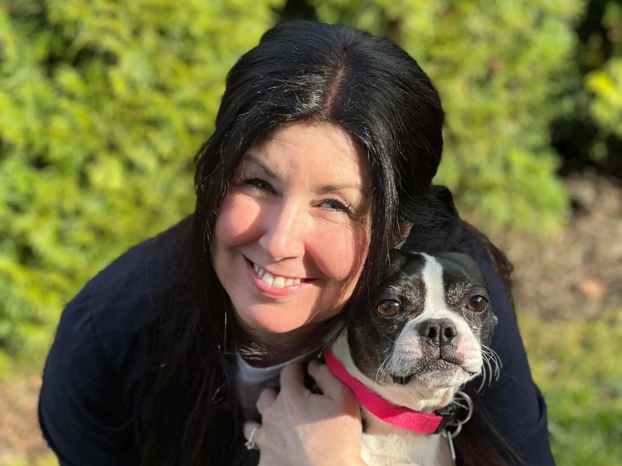 A woman with black hair smiling while holding a black and white puppy with a red collar outdoors on a sunny day.