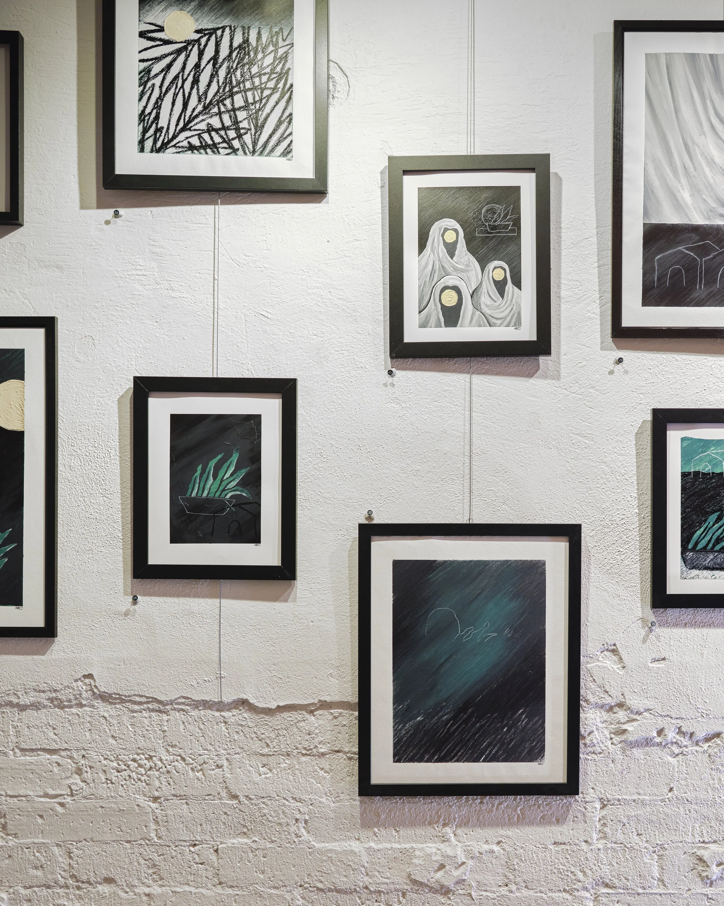 Gallery wall displaying various abstract and illustrative artworks framed in black, green, and white frames, on a textured white wall.