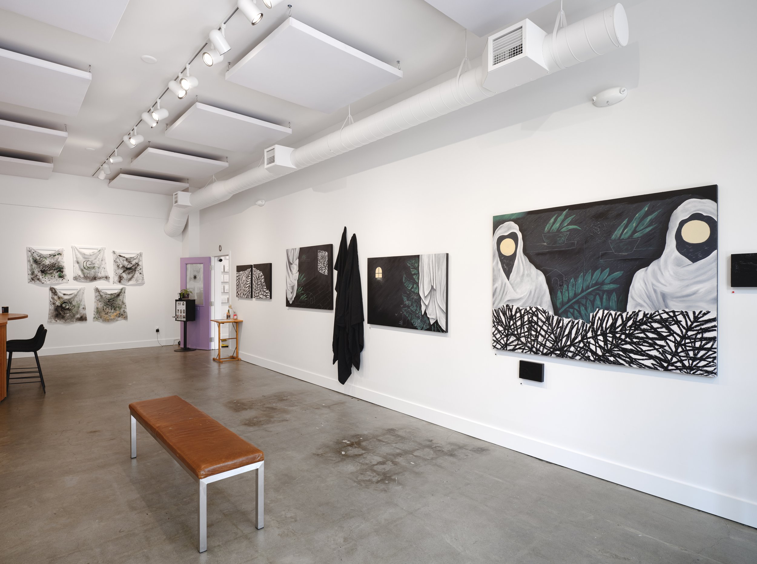 Art gallery interior with paintings on white walls, a bench, and some black coats hanging on a hook. Artwork includes black and white sketches and colorful pieces featuring figures and nature elements.