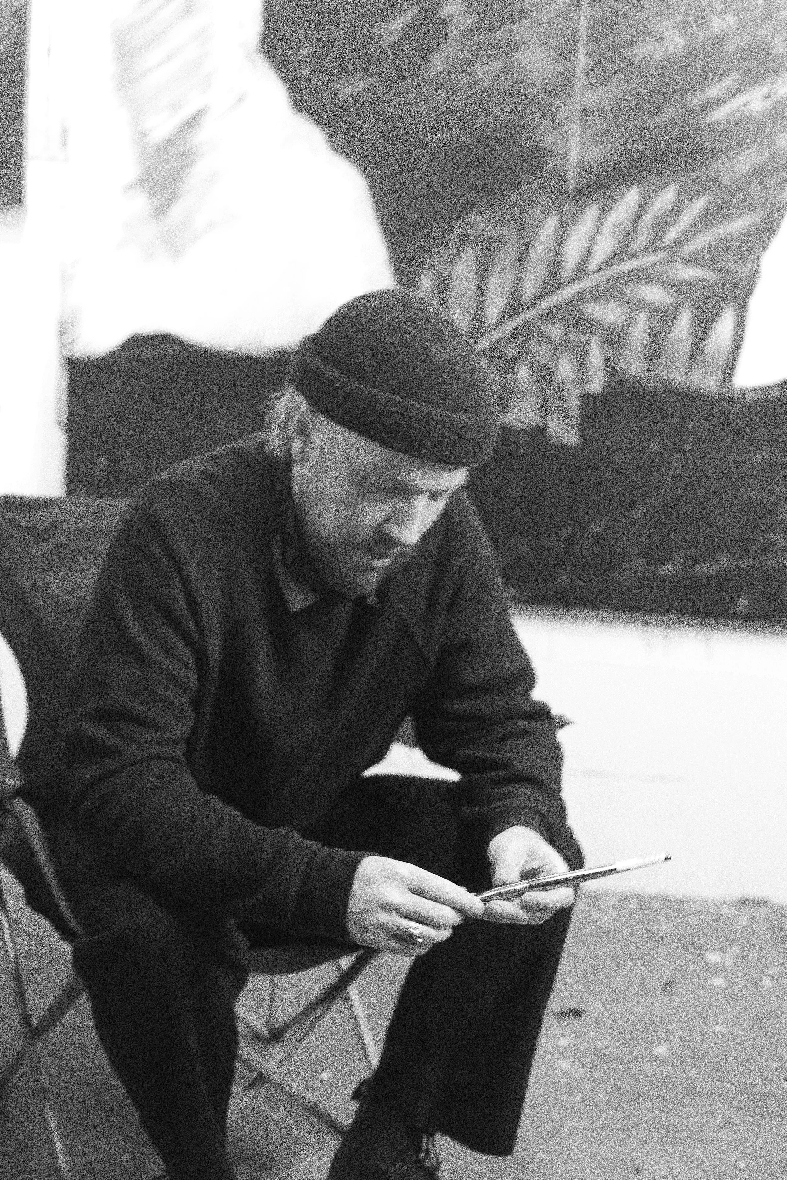 A man with a beard and long hair, wearing a black beanie and dark clothing, is sitting on a folding chair and looking at his phone. There is a large painting of a leaf in the background.