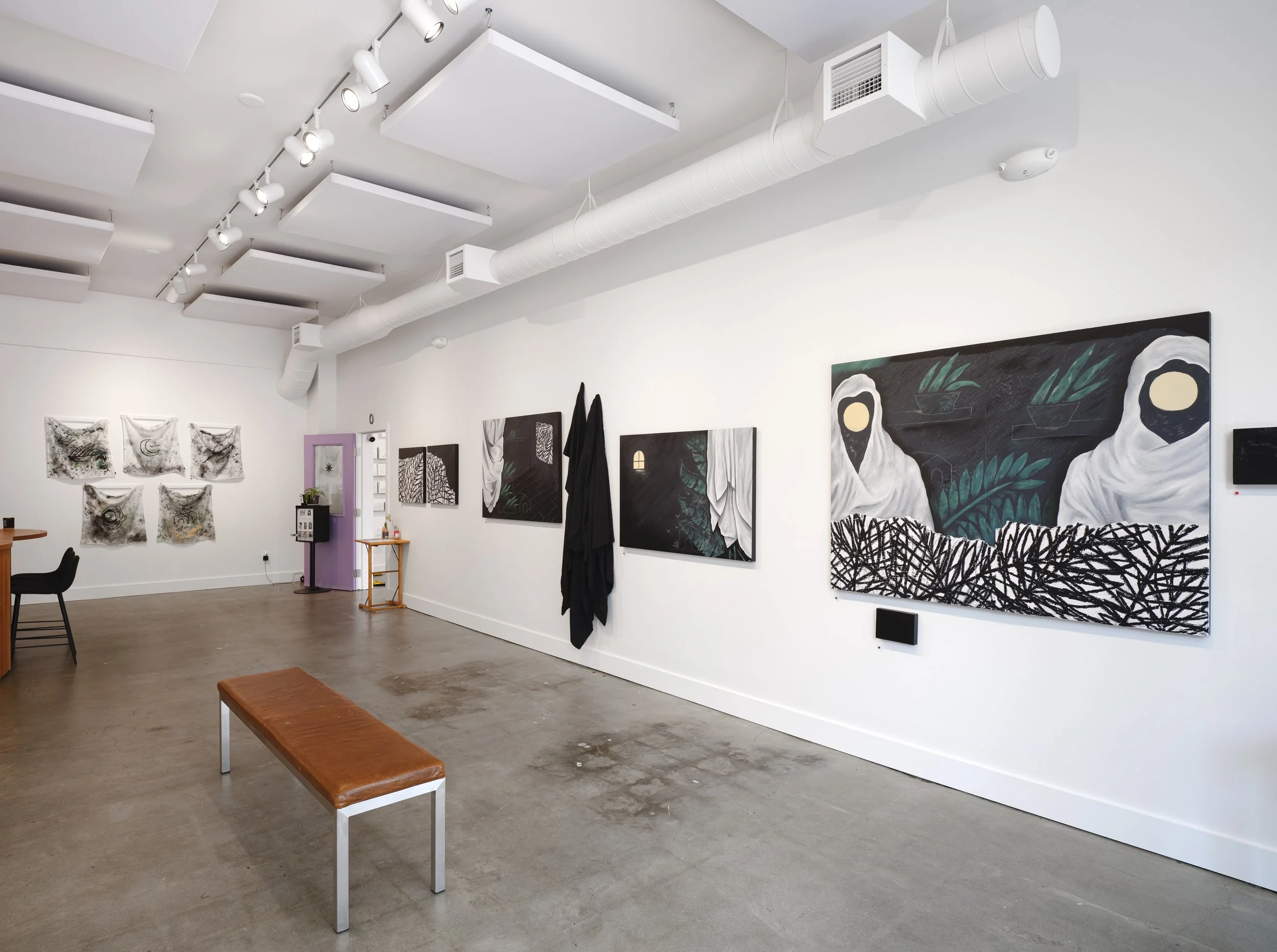 Contemporary art gallery with white walls and ceiling, featuring multiple paintings and textile art pieces, with a bench in the foreground and a small table near a lavender door.