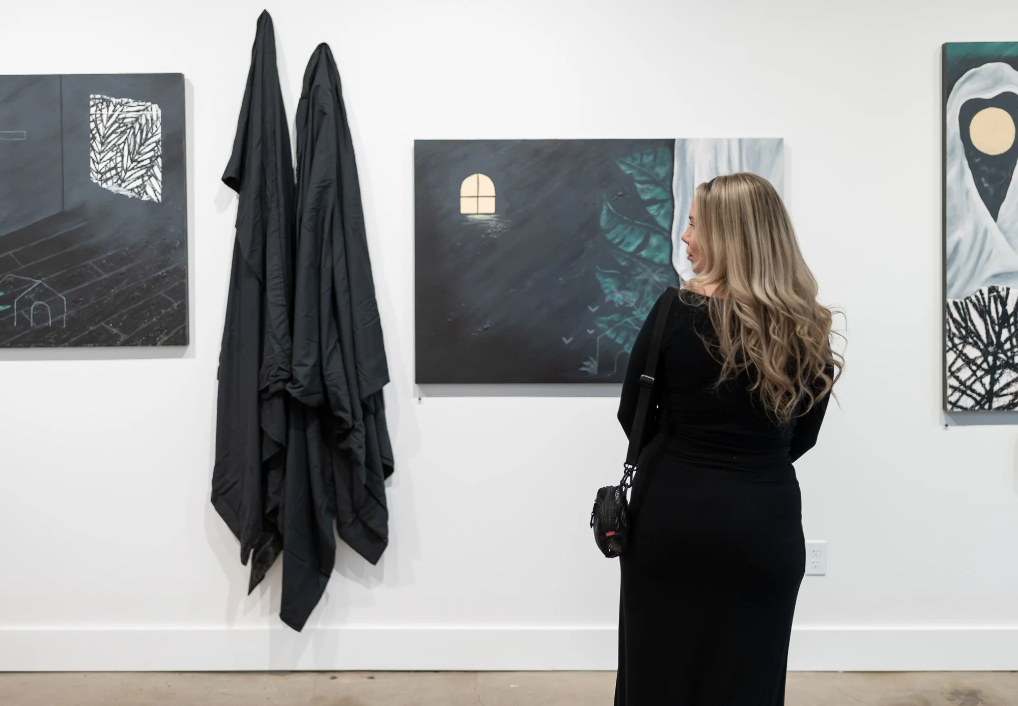 A woman with long blonde hair wearing a black dress is looking at artwork in an art gallery. There are three paintings on the wall, and a black coat hanging on the wall to her left.
