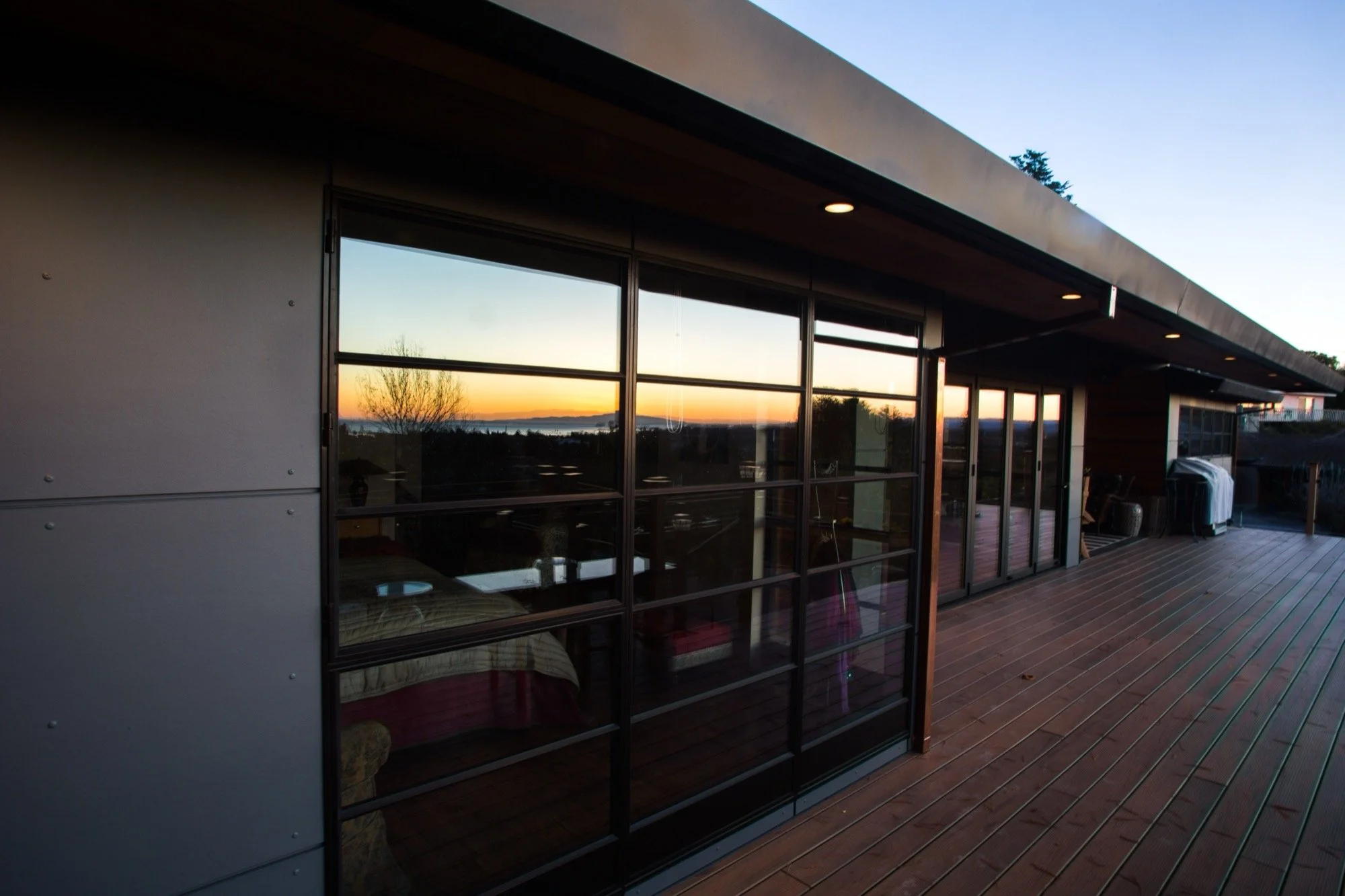 A modern house balcony with wooden flooring, large glass sliding doors reflecting a sunset sunset, and a view of trees and water in the distance.