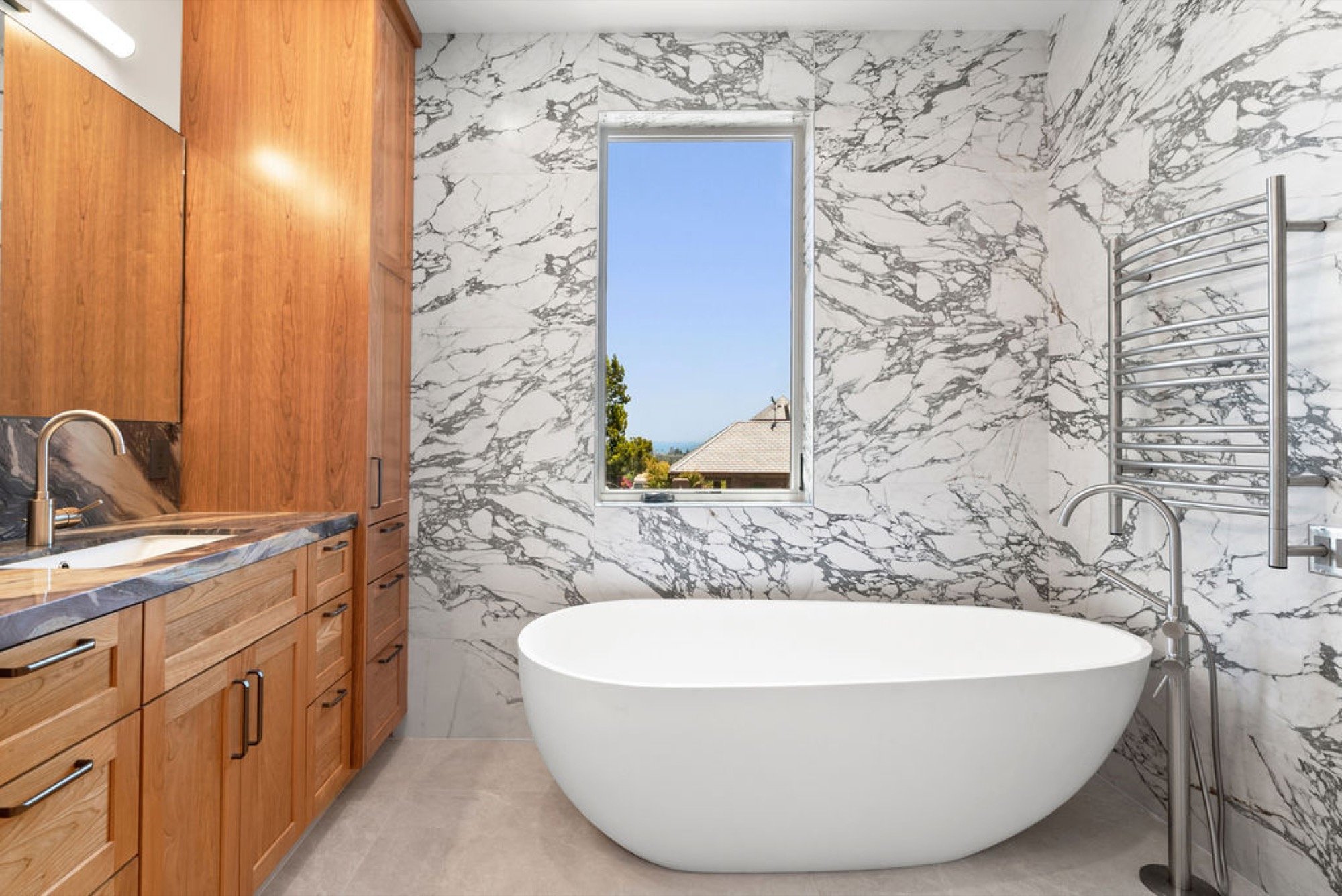 Modern bathroom with a white freestanding bathtub, marble walls, wooden cabinetry with a sink, and a window showing a house and trees outside.