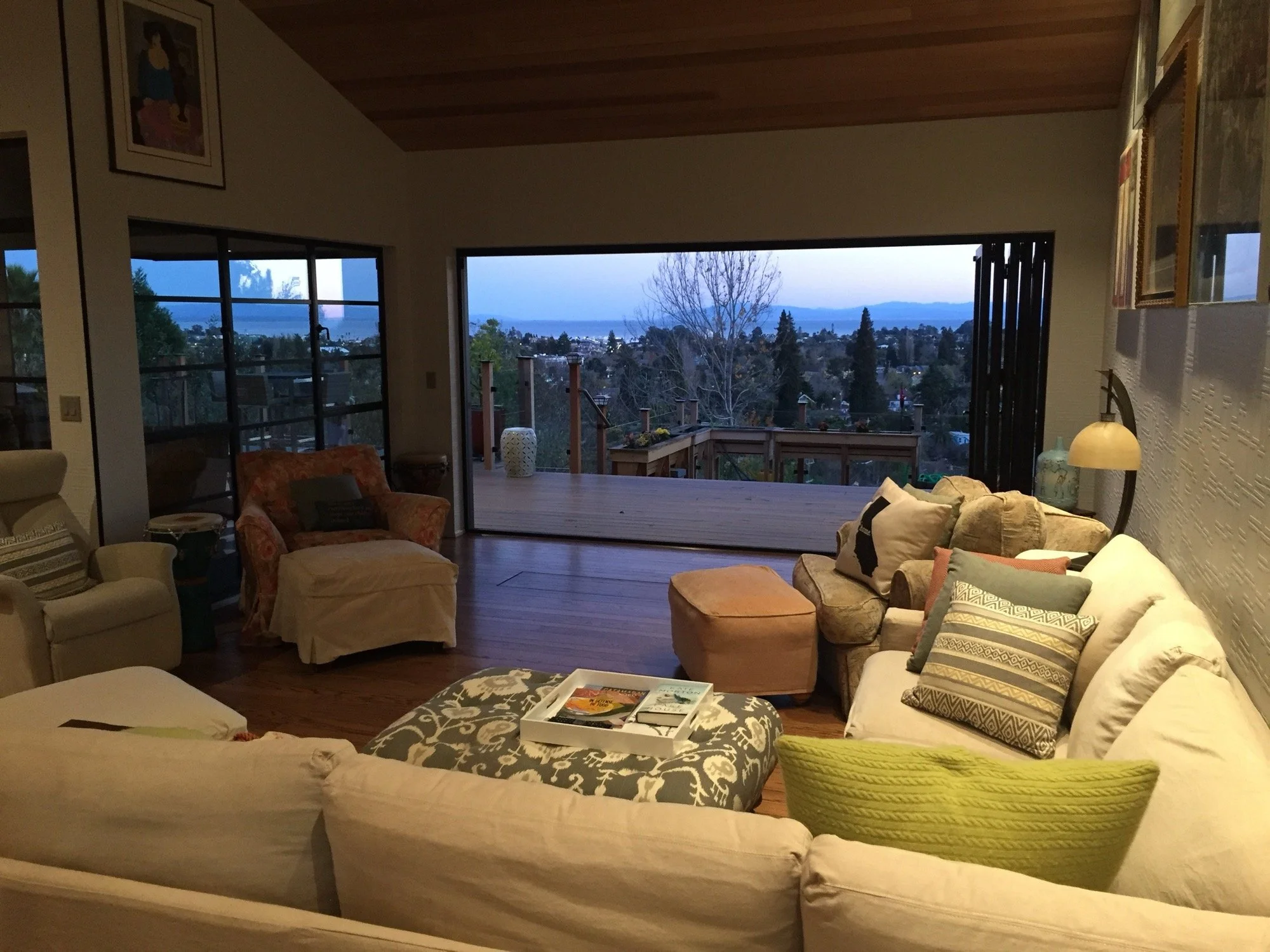 Living room with a large white sectional sofa, cushions, and an ottoman, facing an open sliding door leading to a deck with a view of trees and a distant landscape at dusk.