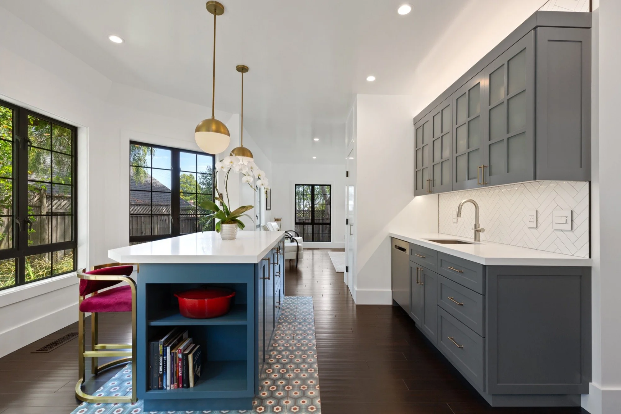 Modern kitchen with gray cabinets, white countertops, and a blue island with gold fixtures. Large windows with black frames, hardwood floors, and decorative pendant lights. Visible dining area in the background.